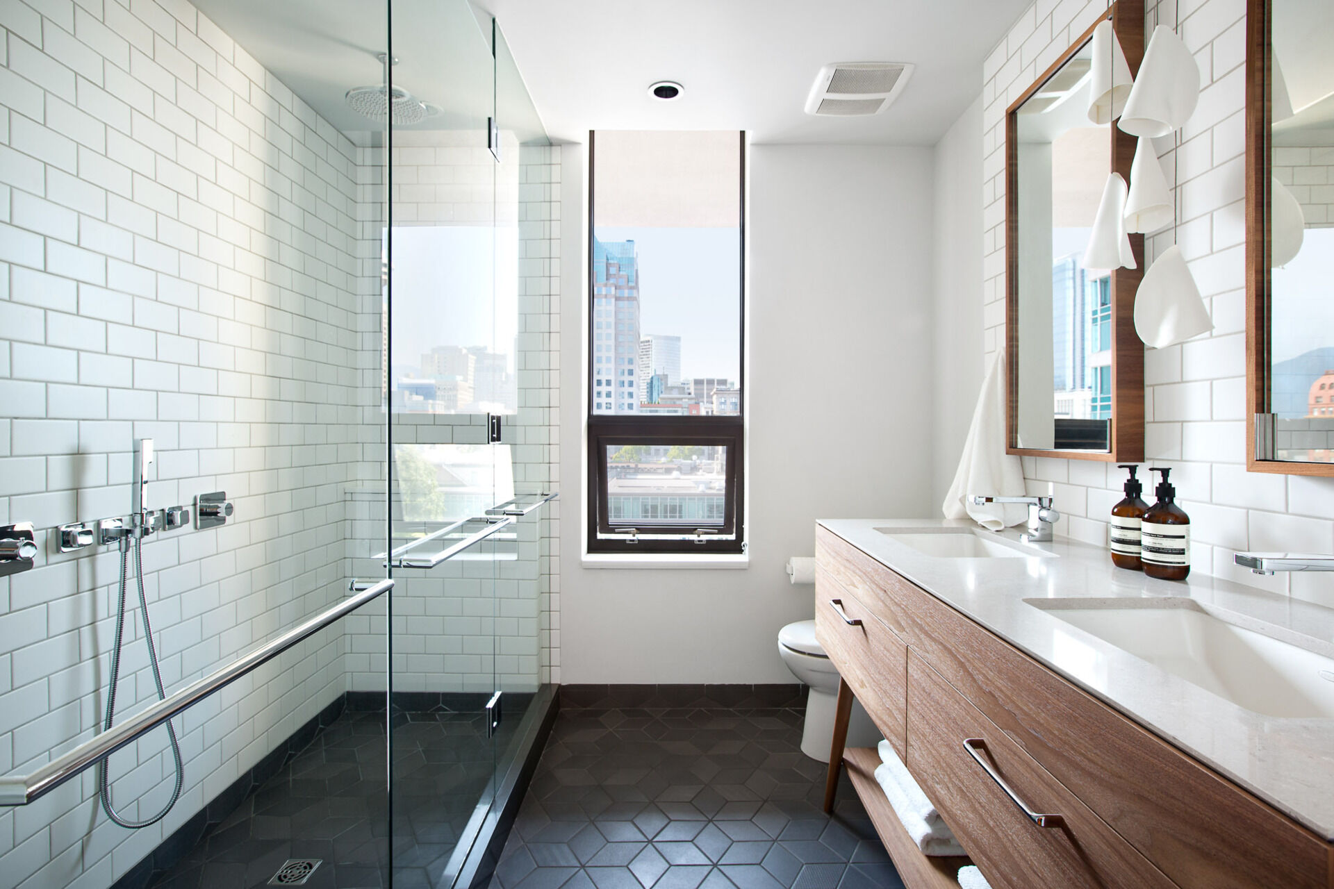 In the bathroom, white subway tiles climb the walls while dark grey geometric floor tiles anchor the space. A wood vanity with dual sinks mirrors the materials used below, giving the entire loft a sense of connection and flow.