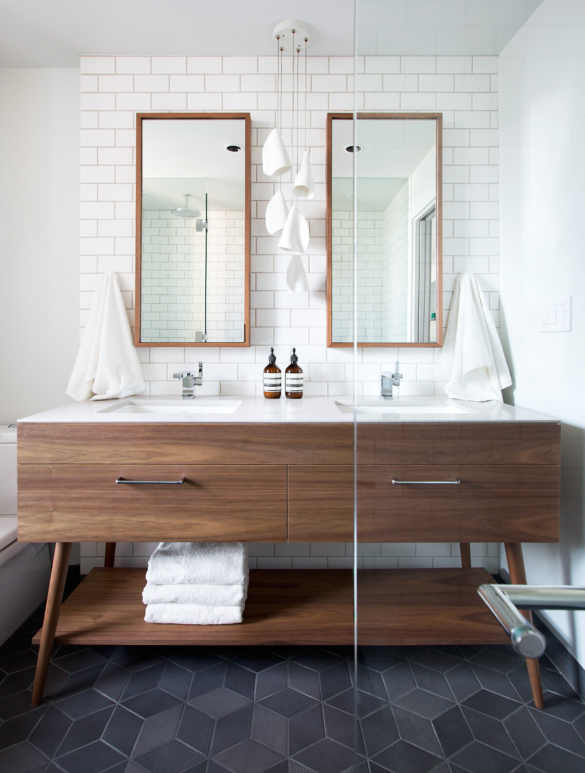 In the bathroom, white subway tiles climb the walls while dark grey geometric floor tiles anchor the space. A wood vanity with dual sinks mirrors the materials used below, giving the entire loft a sense of connection and flow.