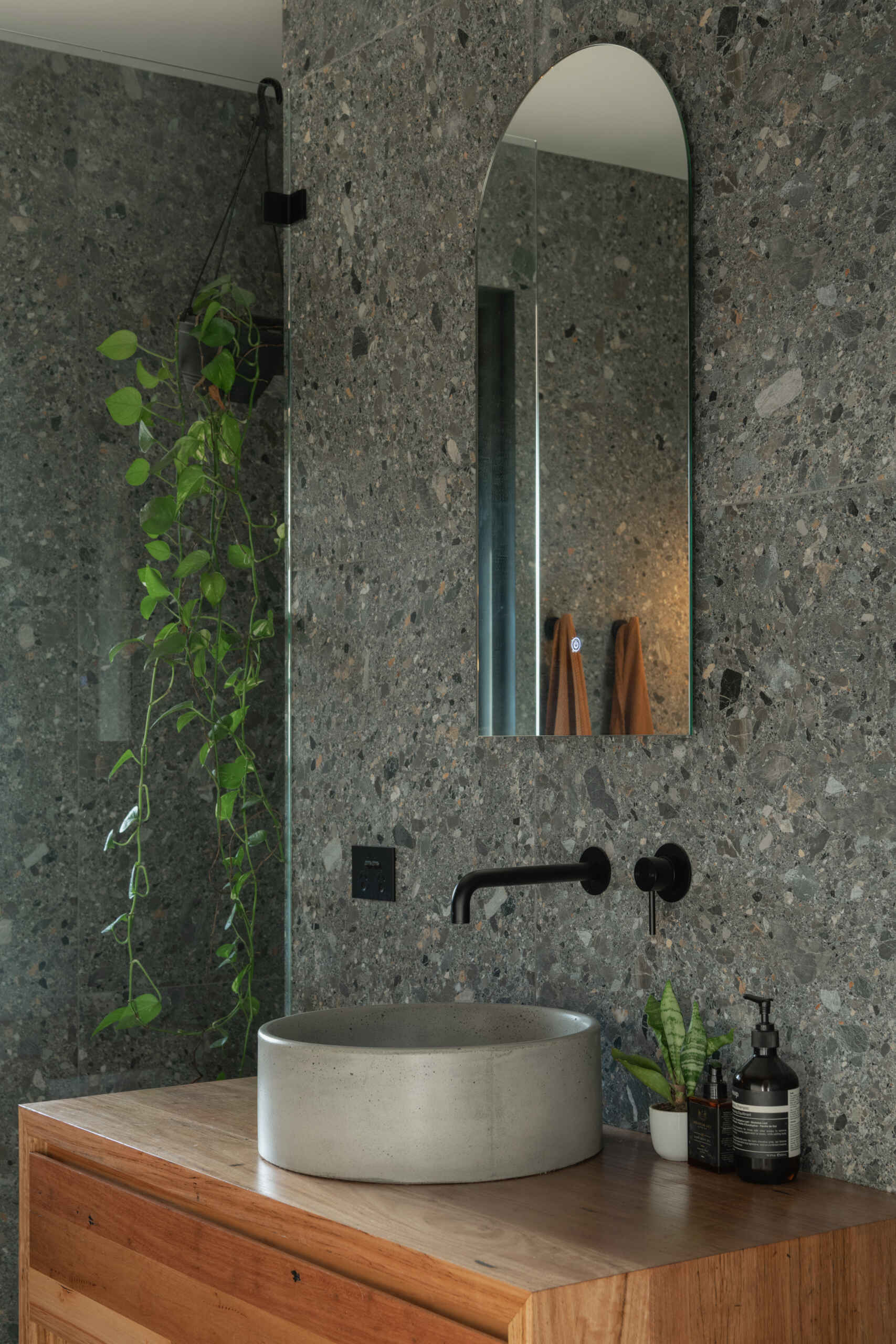 A mix of concrete, timber, and matte black fixtures defines the bathroom’s refined minimalism. The arched mirror and greenery introduce softness, transforming the space into a serene retreat.