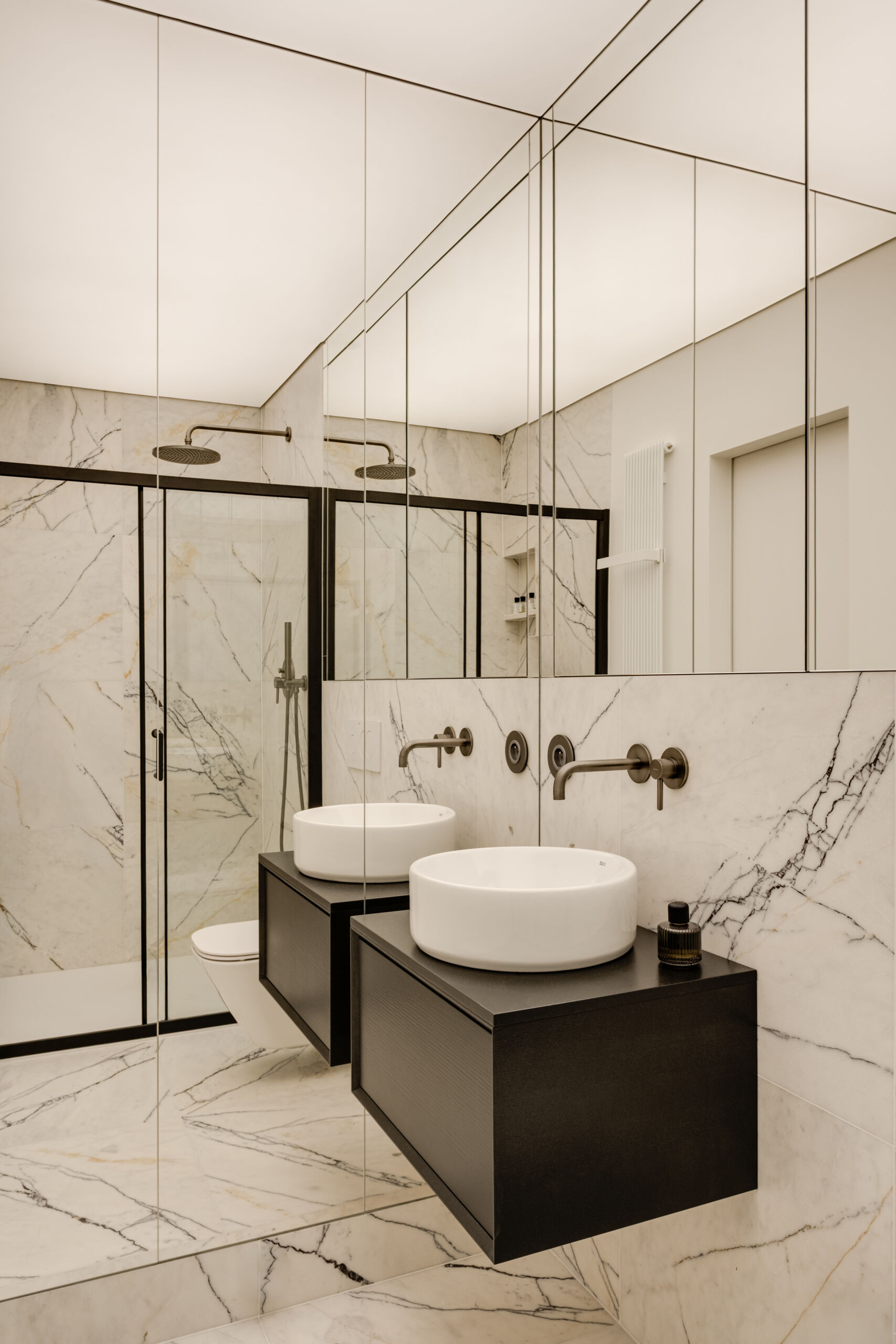 A mirrored cube hides an entire bathroom and wardrobe in plain sight. Its reflections expand the small apartment, while inside, white Calacatta Lilac marble and soft backlighting make the space feel impossibly bright and tall.