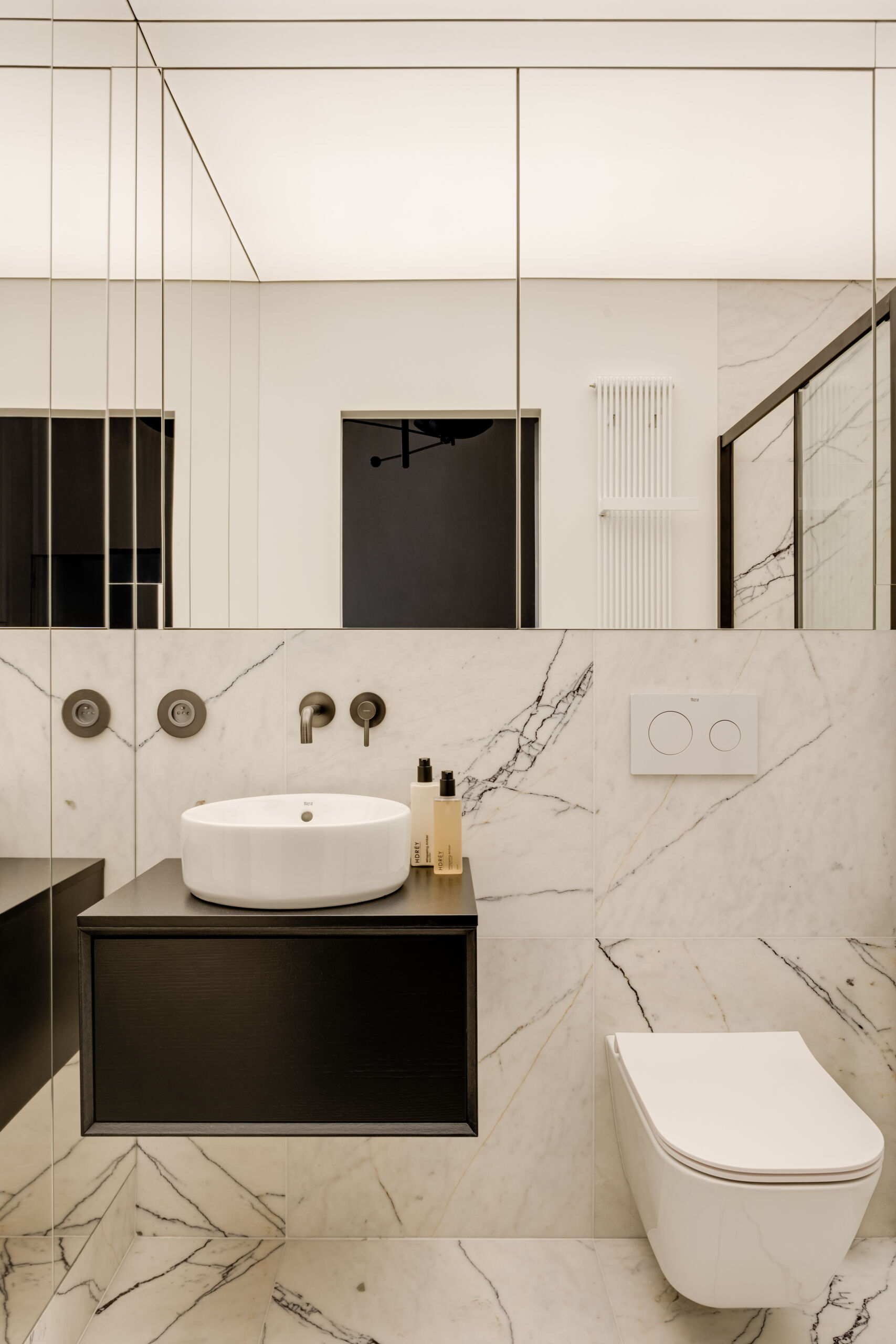 A mirrored cube hides an entire bathroom and wardrobe in plain sight. Its reflections expand the small apartment, while inside, white Calacatta Lilac marble and soft backlighting make the space feel impossibly bright and tall.