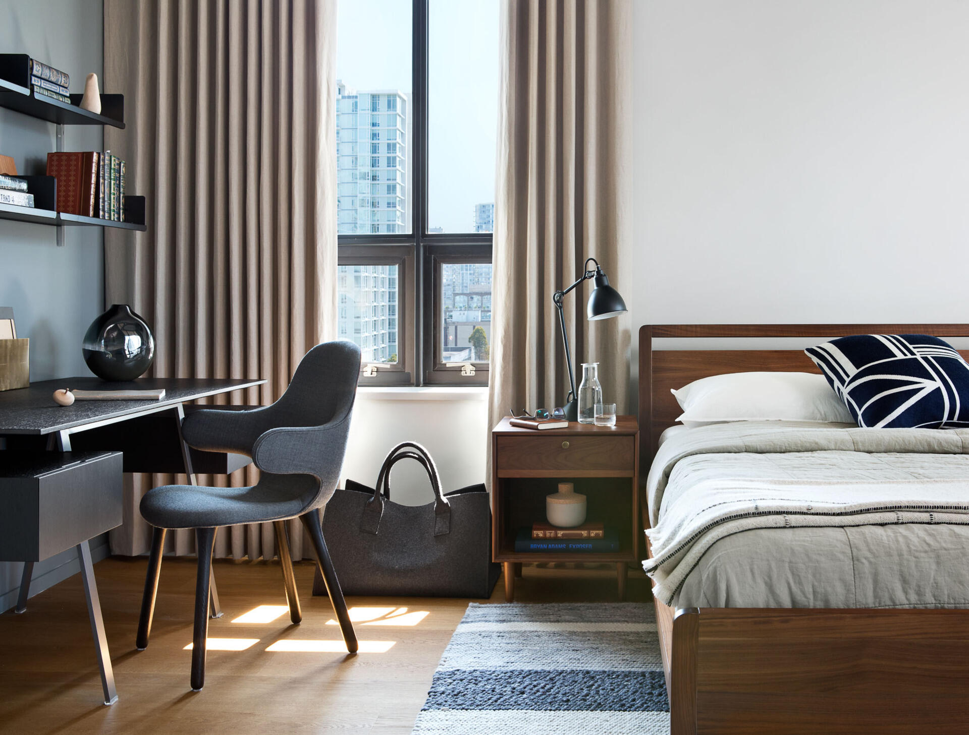 A simple wood bed frame and side tables feel tactile and grounded, while a small workspace with open shelving tucks neatly beside the window. Every surface serves a purpose without overwhelming the calm.