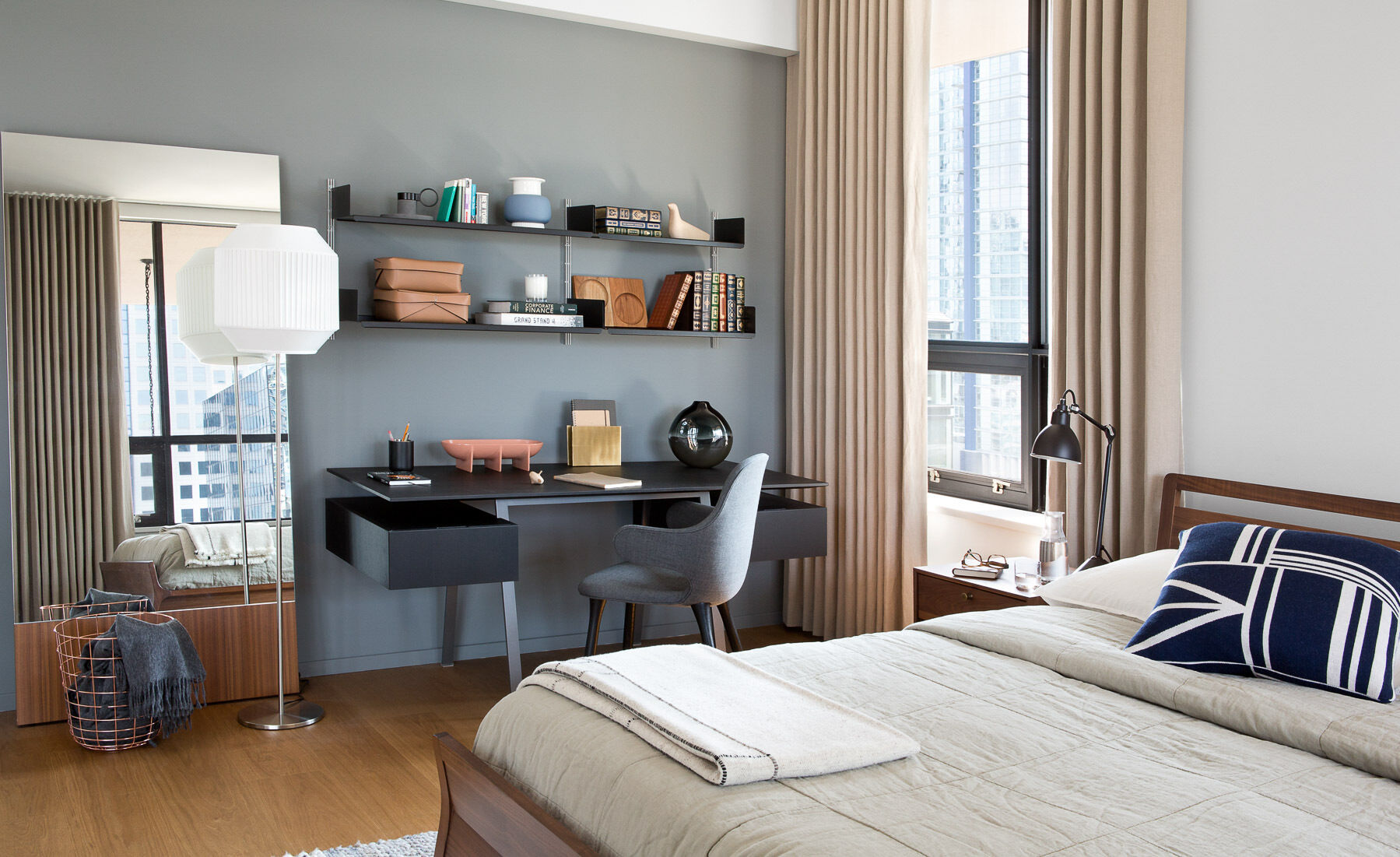 A simple wood bed frame and side tables feel tactile and grounded, while a small workspace with open shelving tucks neatly beside the window. Every surface serves a purpose without overwhelming the calm.