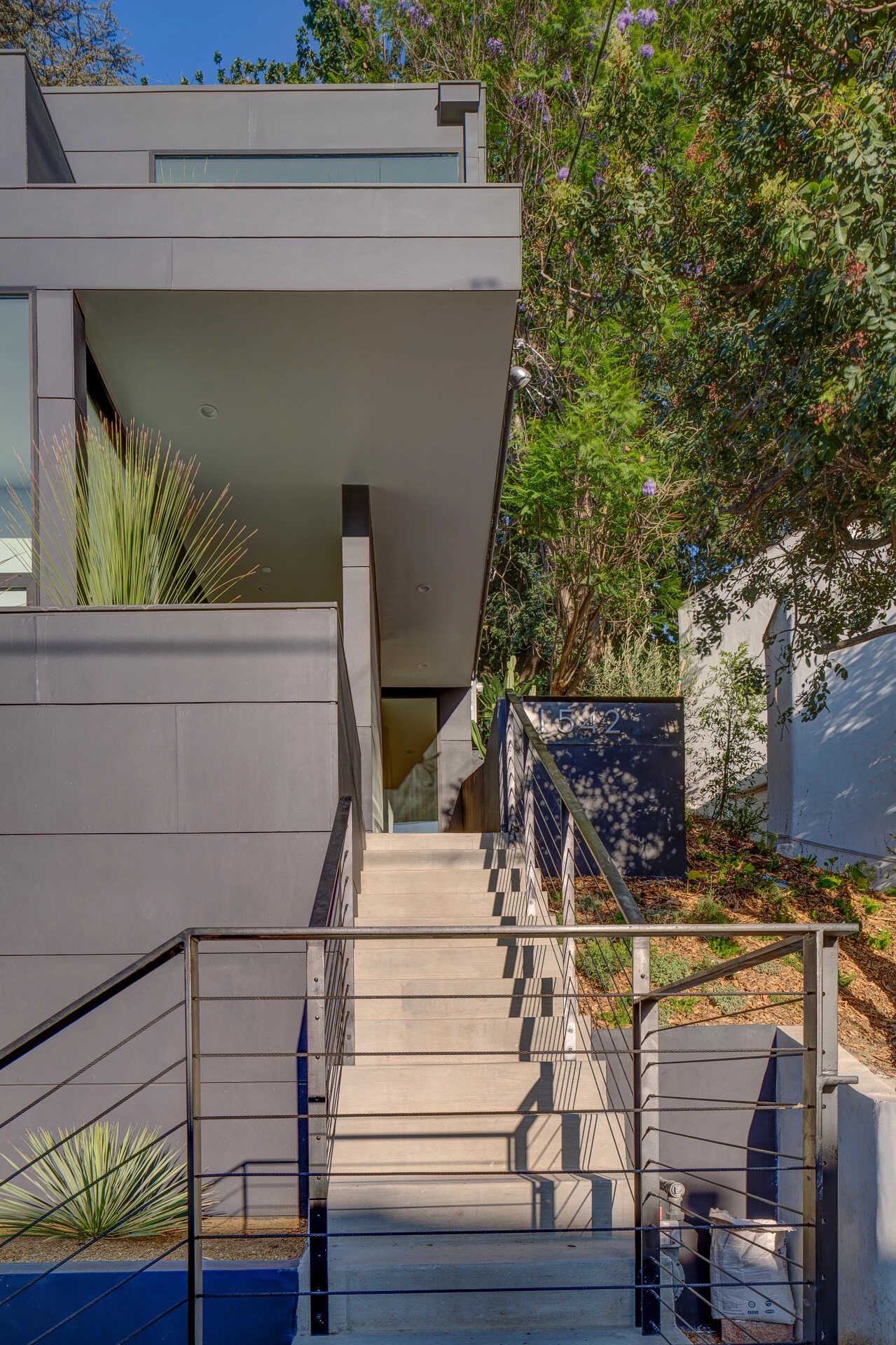 From the street, concrete steps rise sharply along the slope, flanked by a minimalist black handrail that mirrors the home’s bold exterior. At the top, a small covered deck provides a moment of pause, a sheltered landing before entering the house.