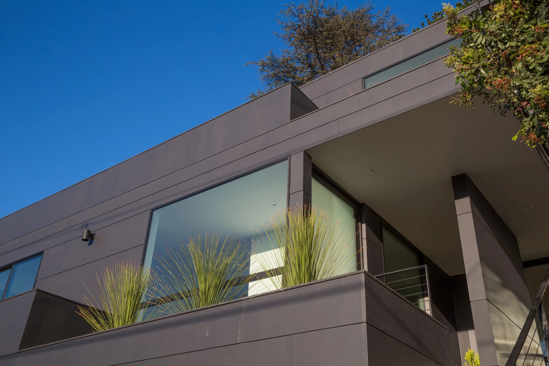 From the street, concrete steps rise sharply along the slope, flanked by a minimalist black handrail that mirrors the home’s bold exterior. At the top, a small covered deck provides a moment of pause, a sheltered landing before entering the house.