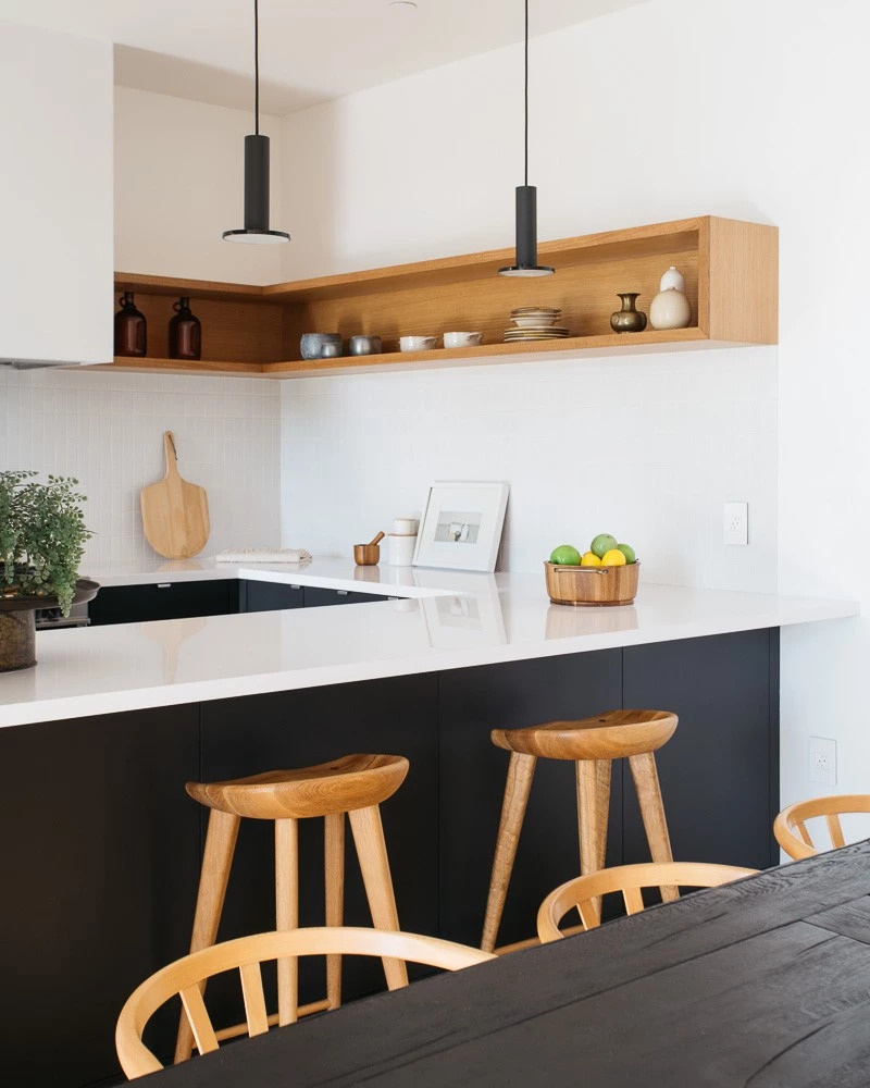In the kitchen, contrast reigns. The upper walls and shelves are bright white and natural wood, while the lower cabinetry is a deep matte black. This balance of light and dark keeps the room feeling fresh yet grounded. The simplicity of the materials highlights the precision of the design, every line and joint perfectly aligned.