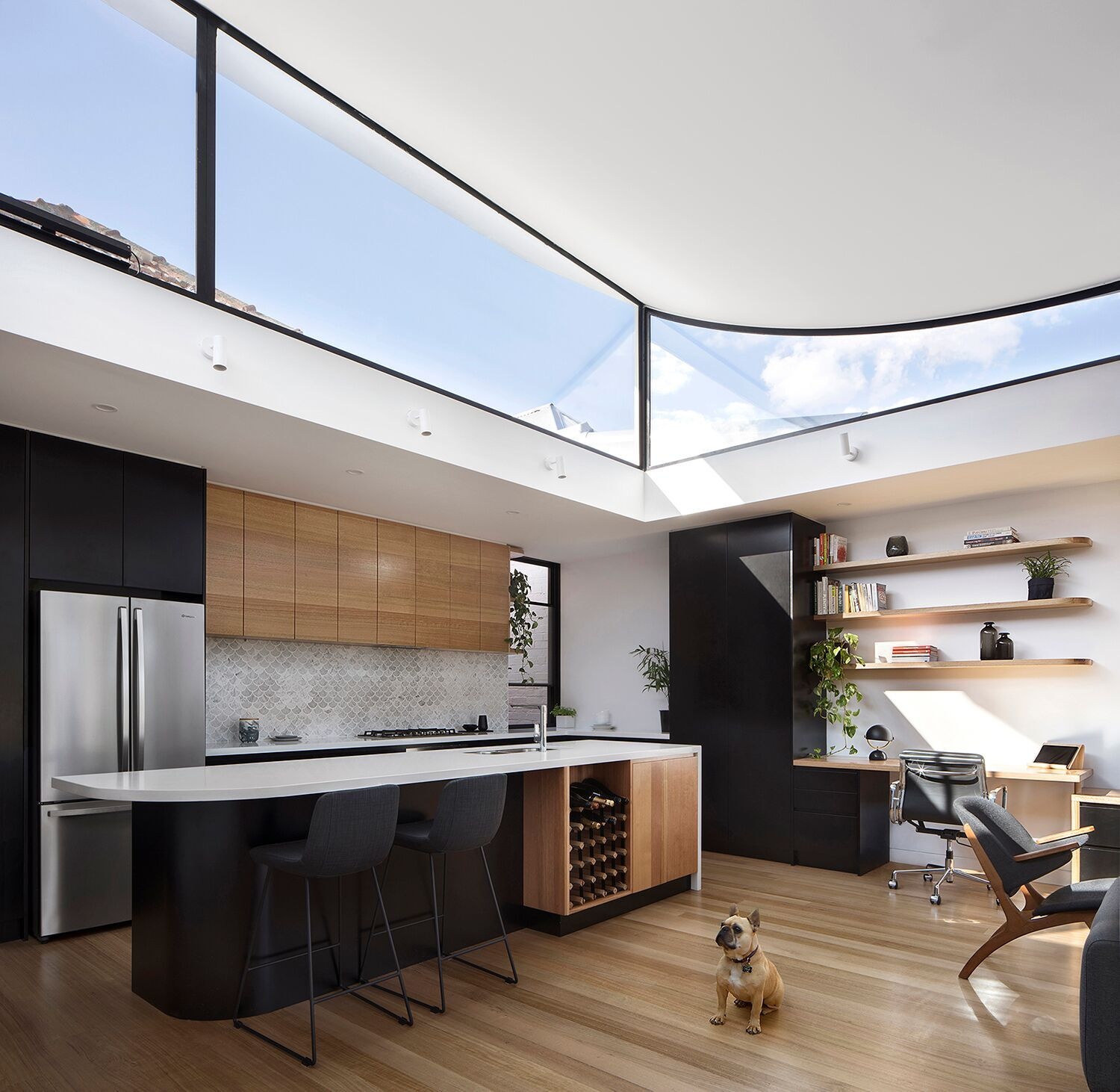 In the kitchen, wood and black cabinetry pair with crisp white counters. A light grey fish scale tile adds texture behind the workspace. A built-in wine rack is tucked into the kitchen island, while curved shelves at the edge of the upper cabinets tie the design back to the office joinery. The effect is continuous and considered, uniting every corner of the open plan.