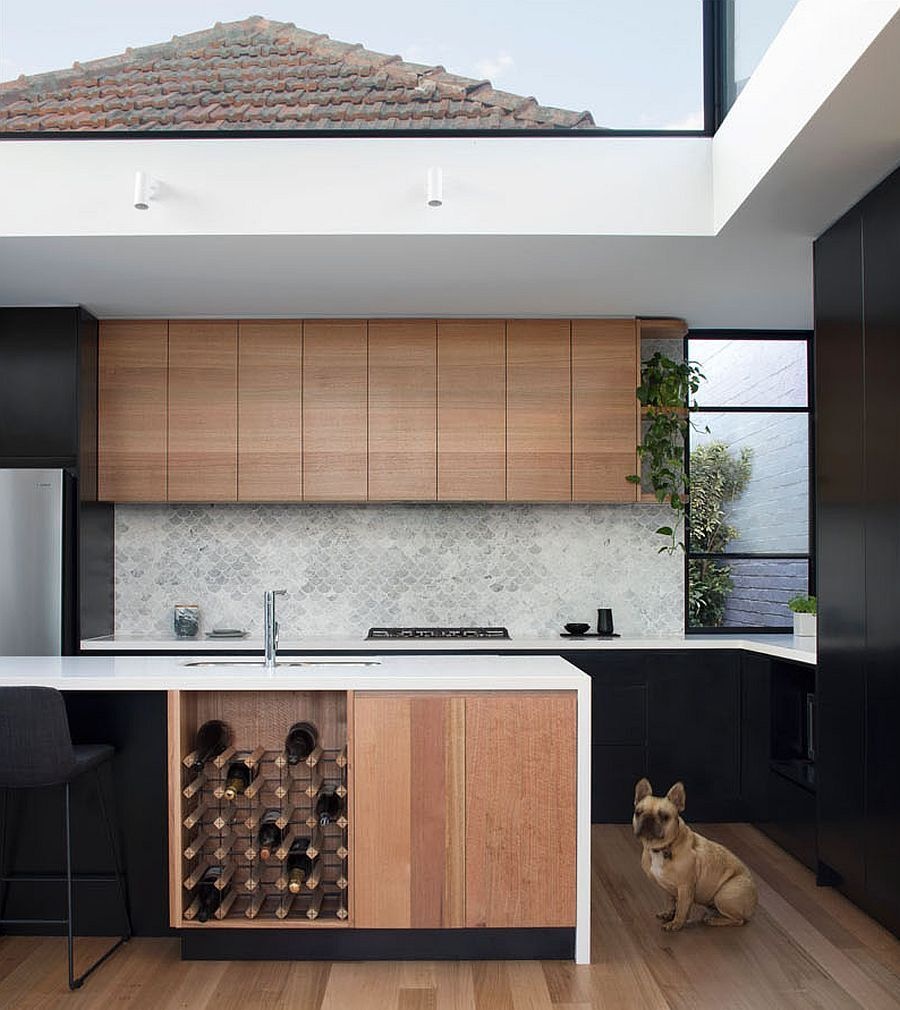 In the kitchen, wood and black cabinetry pair with crisp white counters. A light grey fish scale tile adds texture behind the workspace. A built-in wine rack is tucked into the kitchen island, while curved shelves at the edge of the upper cabinets tie the design back to the office joinery. The effect is continuous and considered, uniting every corner of the open plan.
