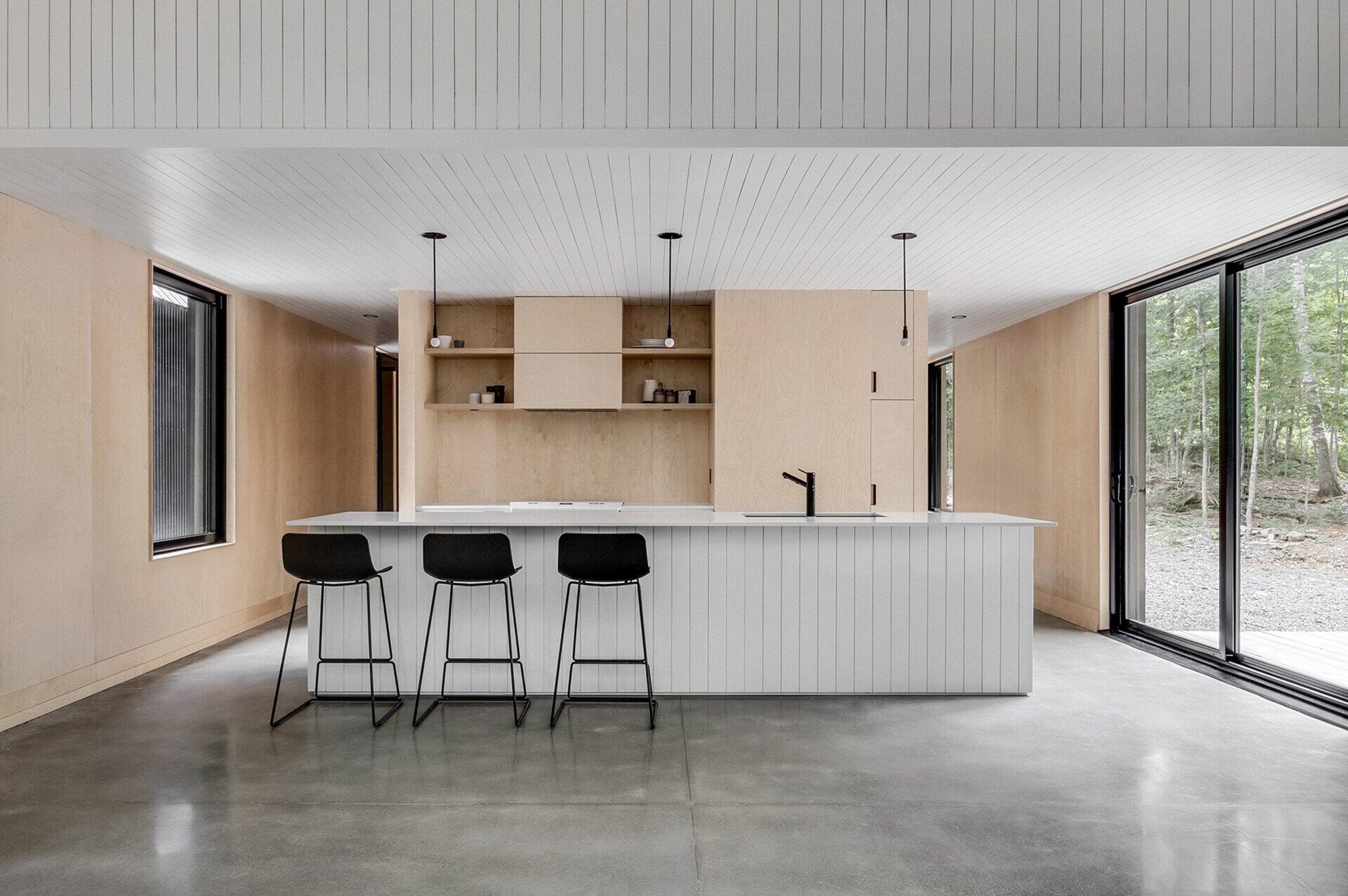 A bright, understated kitchen with polished concrete floors and light wood finishes connects seamlessly to the outdoors through glass doors.