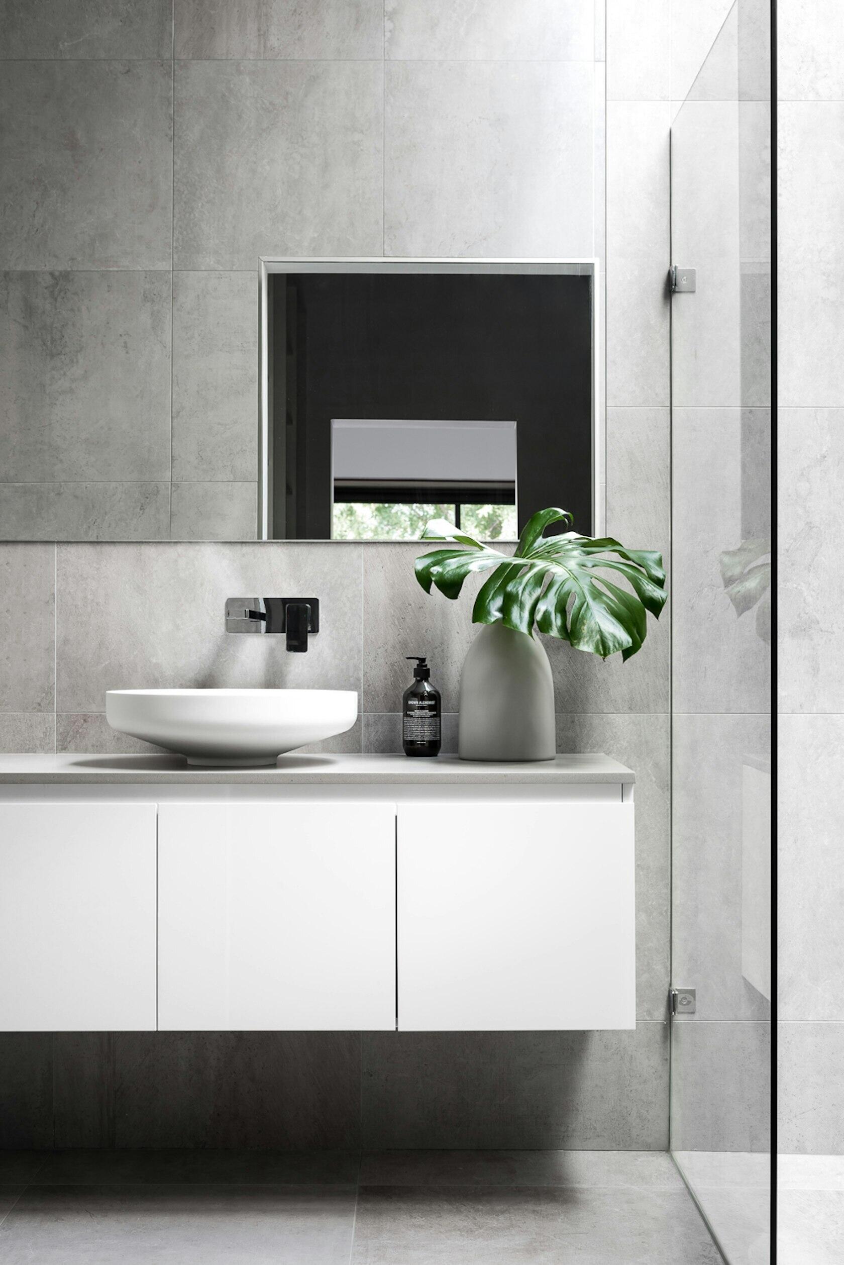 In the primary bathroom, large grey tiles wrap the walls and floors, while a skylight fills the room with daylight. A glass partition defines the shower, keeping the space open and airy beside a simple white floating vanity.