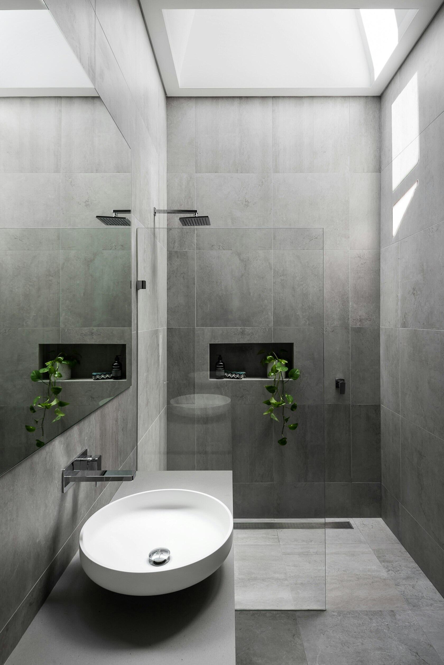 In the primary bathroom, large grey tiles wrap the walls and floors, while a skylight fills the room with daylight. A glass partition defines the shower, keeping the space open and airy beside a simple white floating vanity.
