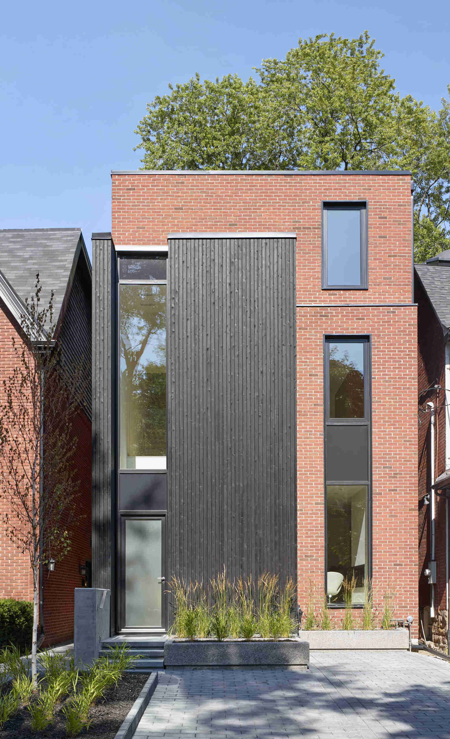 A striking modern home in Toronto’s Summerhill neighborhood by Akb Architects, featuring bold red brick, black-stained cedar, and tall narrow windows that give the facade a graphic, architectural rhythm.