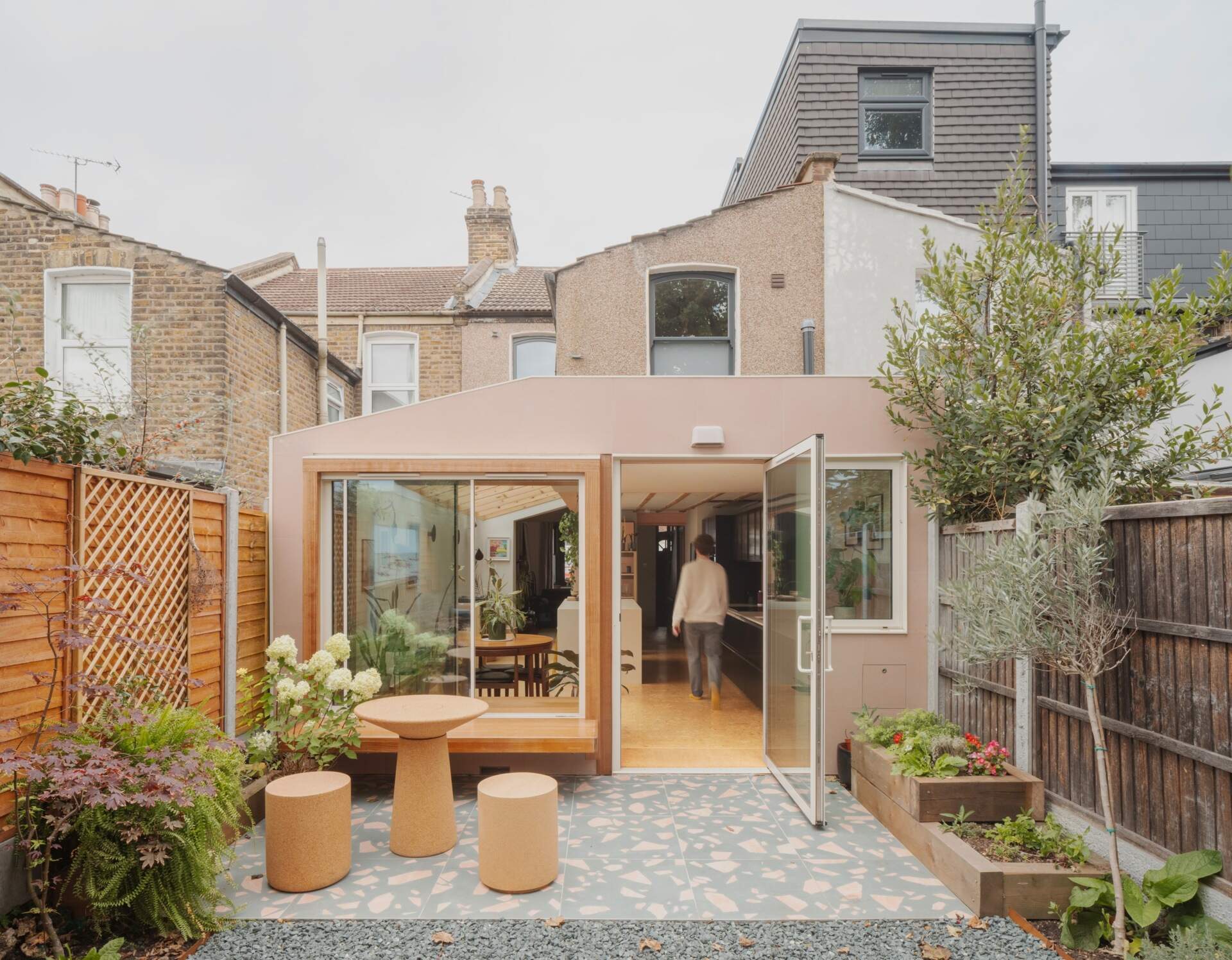 A light-filled garden extension with a new kitchen and dining area, includes pink steel, cork flooring, a pivoting door, and rooflights.
