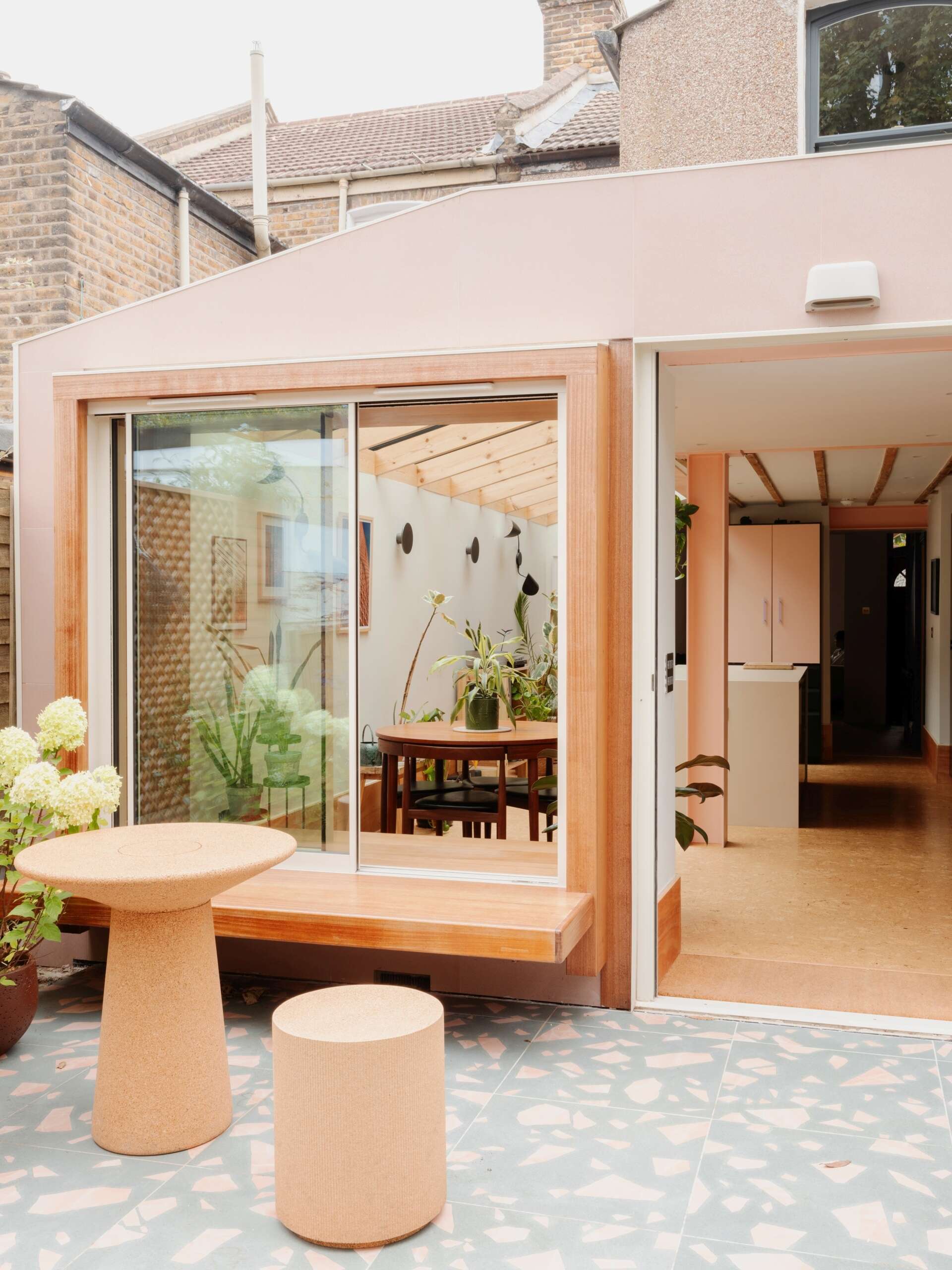 A light-filled garden extension with a new kitchen and dining area, includes pink steel, cork flooring, a pivoting door, and rooflights.
