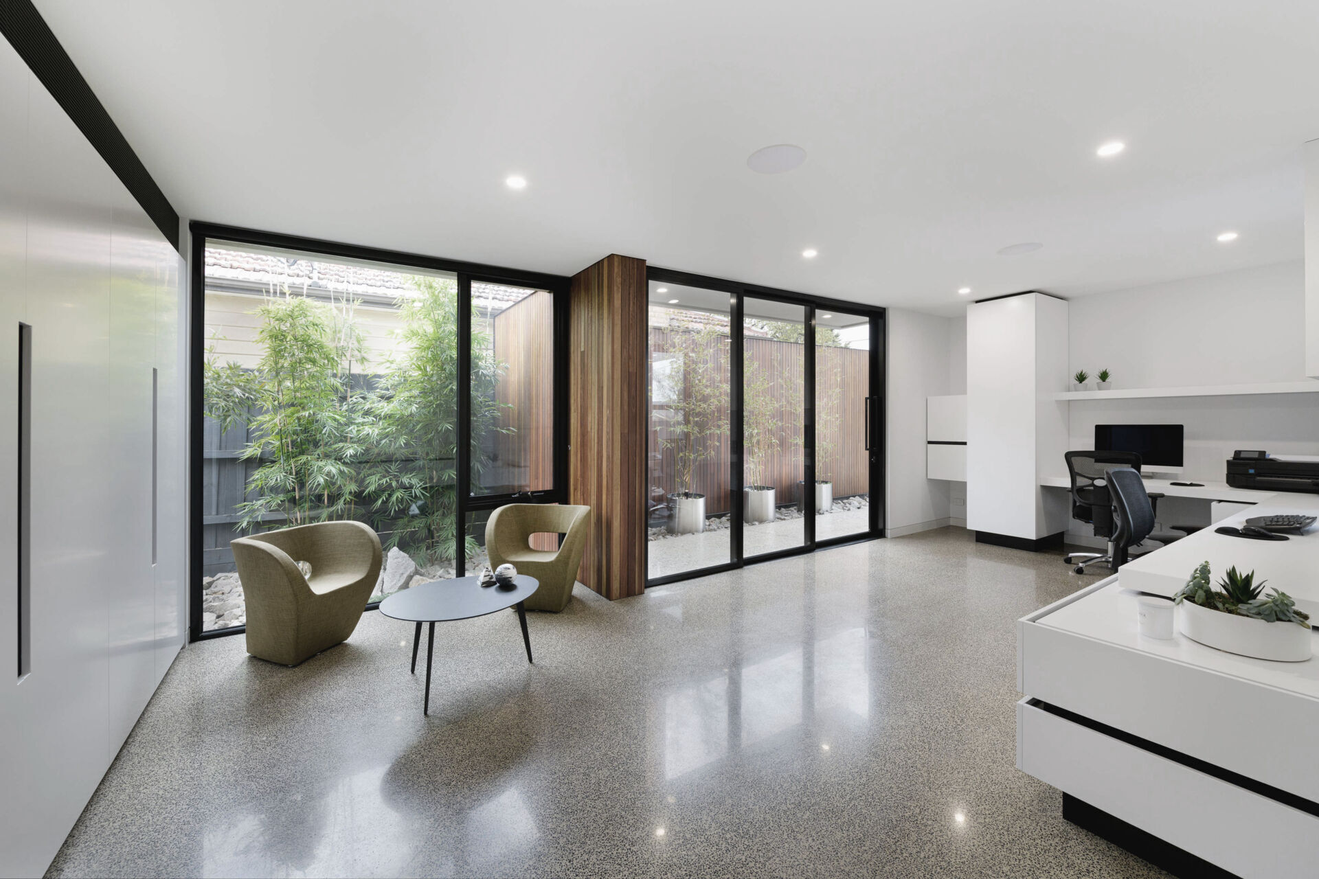 This home office has polished concrete floors reflect soft daylight that filters through full-height glazing, framing views of bamboo and timber screens outside. Minimal white cabinetry and built-in shelving keep the focus on the open, light-filled atmosphere.