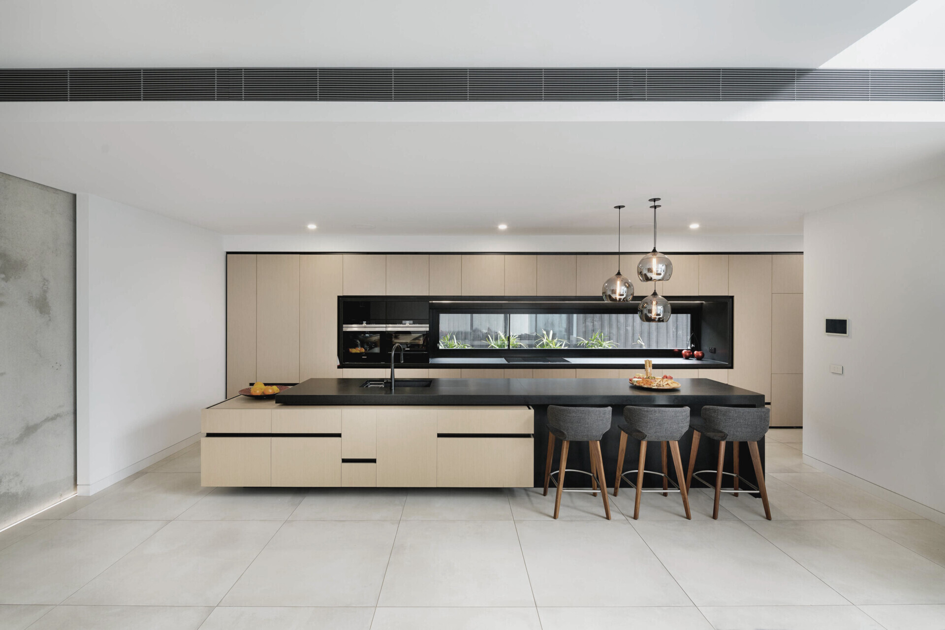 This kitchen continues the home’s language of precision and restraint. A central island anchors the pale cabinetry, creating a dialogue between strength and softness. Integrated appliances and glass pendant lights maintain a sleek, uninterrupted line, while a horizontal window introduces natural light and a glimpse of greenery.