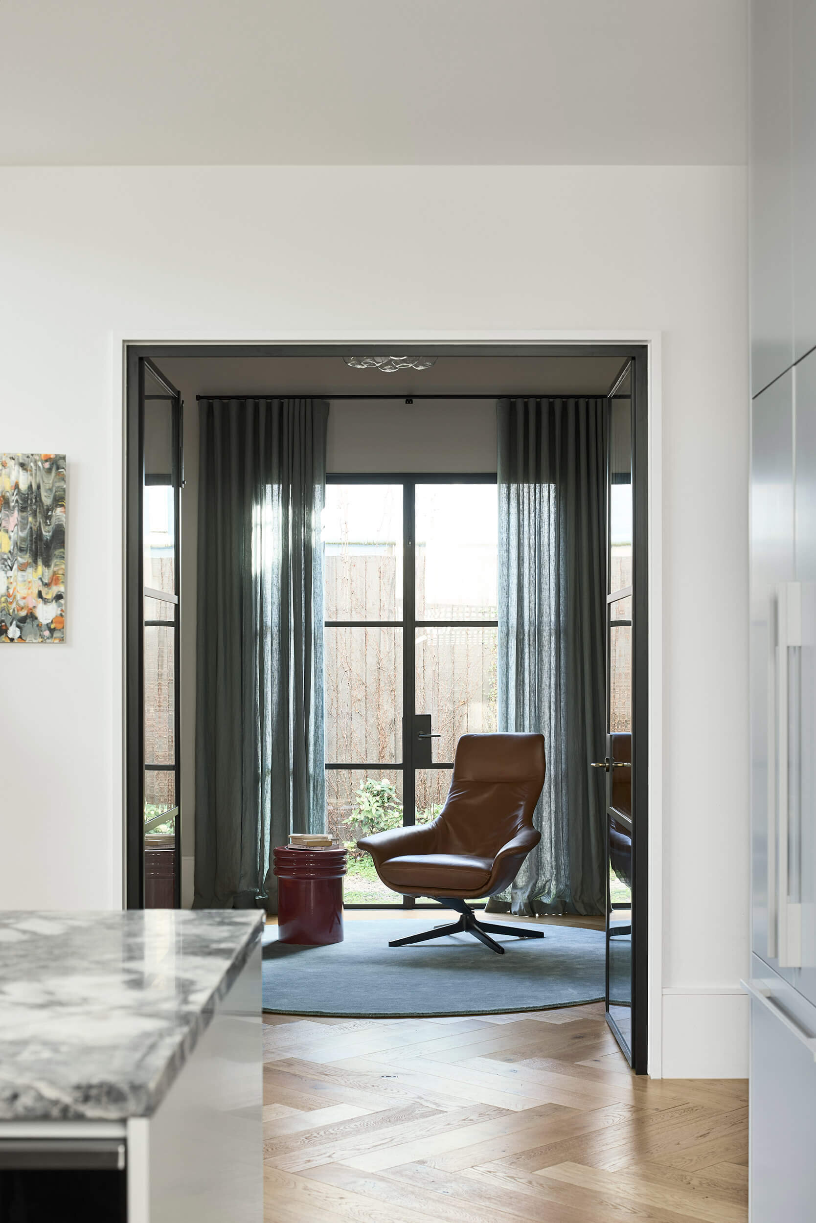 A glass-framed doorway leads into a small yet refined home office where sheer curtains diffuse the brightness, balancing privacy with openness.