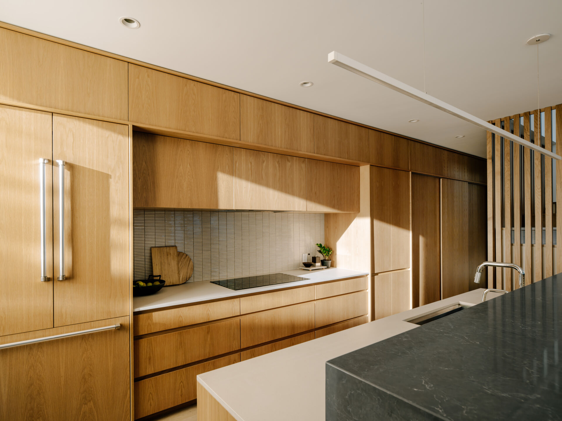 Inside, the kitchen, dining, and living spaces flow along the rear yard. A covered “engawa” porch hovers above the ground, inviting sitting and connecting indoor and outdoor life. Custom white oak built-ins maximize storage and function, including a bench integrated into the kitchen island for casual dining.