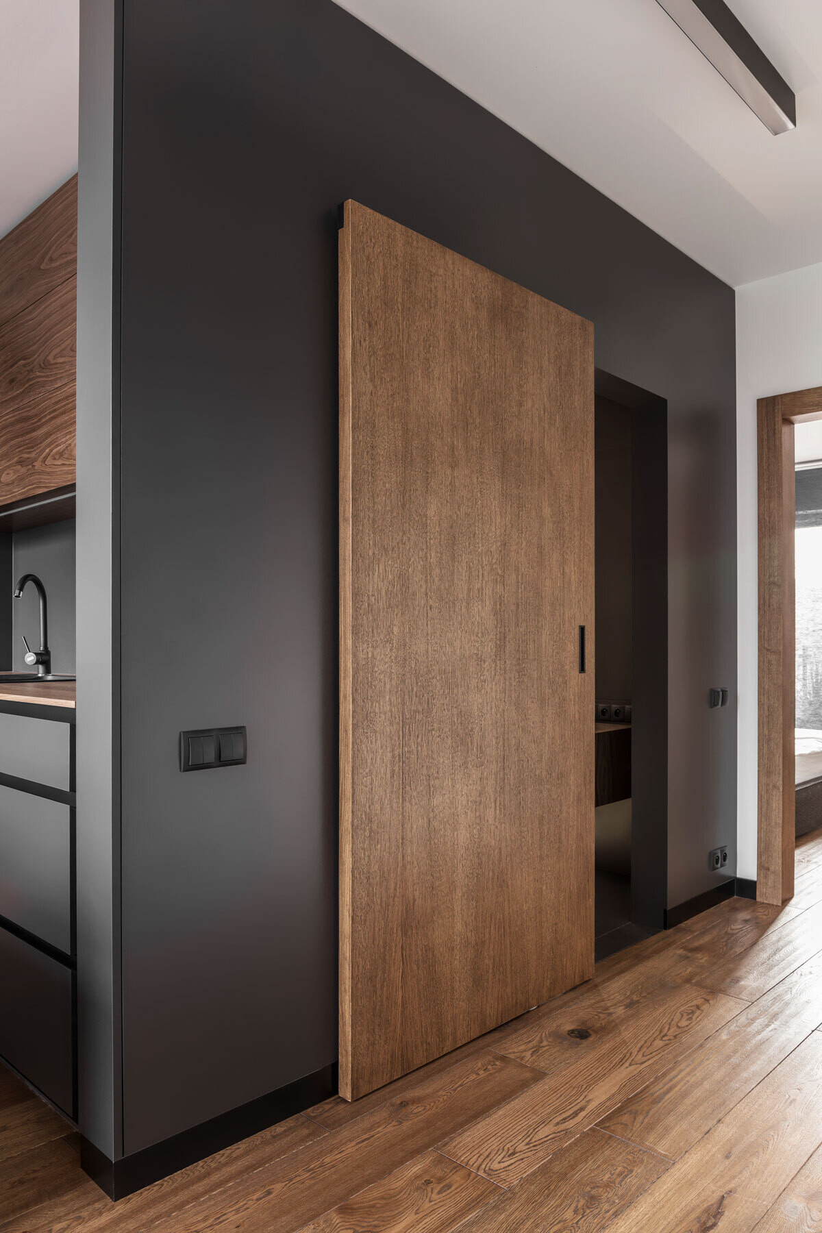 This bathroom hides a surprising layer of comfort. A sliding wooden door opens to reveal a space wrapped in deep tones, with soft floor lighting casting a subtle glow. White tiles and mirrors amplify the light, creating balance and calm.