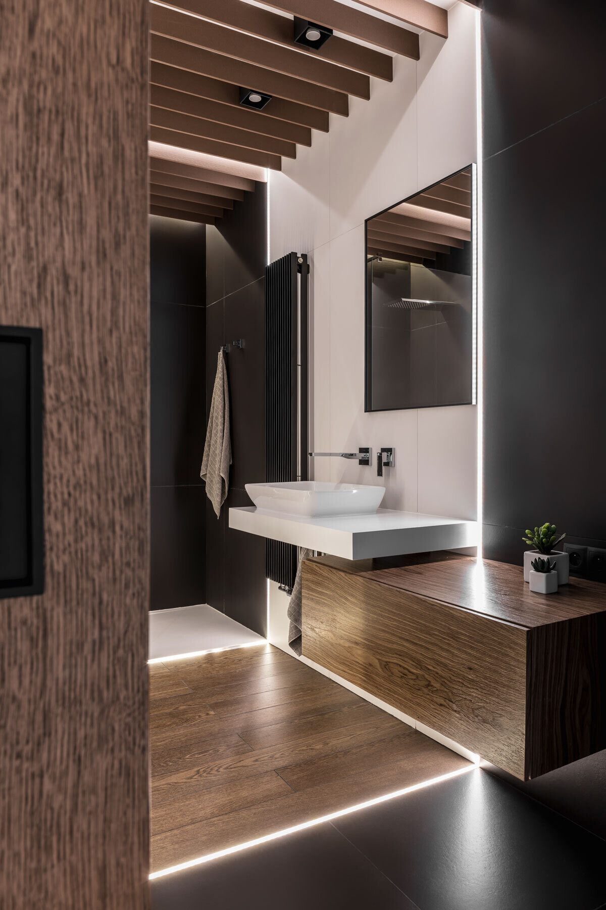 This bathroom hides a surprising layer of comfort. A sliding wooden door opens to reveal a space wrapped in deep tones, with soft floor lighting casting a subtle glow. White tiles and mirrors amplify the light, creating balance and calm.
