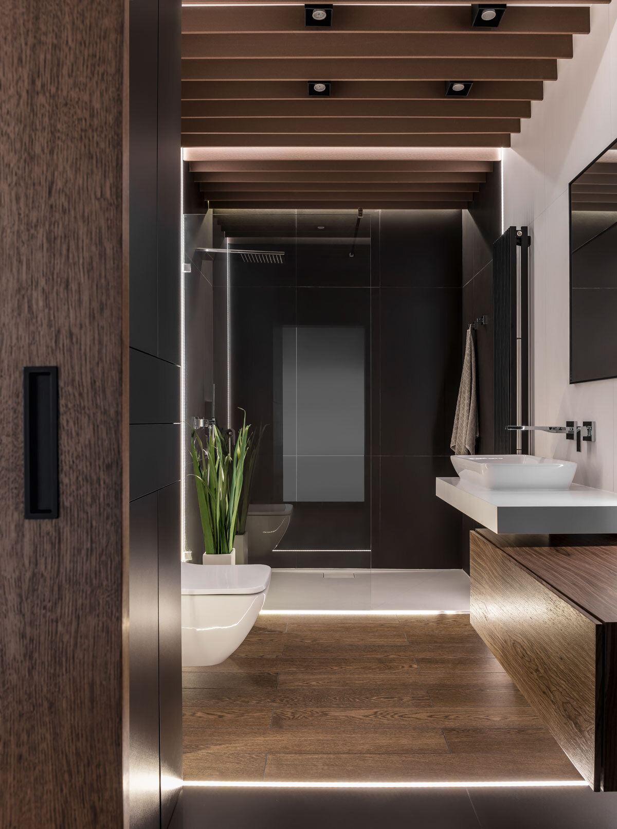 This bathroom hides a surprising layer of comfort. A sliding wooden door opens to reveal a space wrapped in deep tones, with soft floor lighting casting a subtle glow. White tiles and mirrors amplify the light, creating balance and calm.