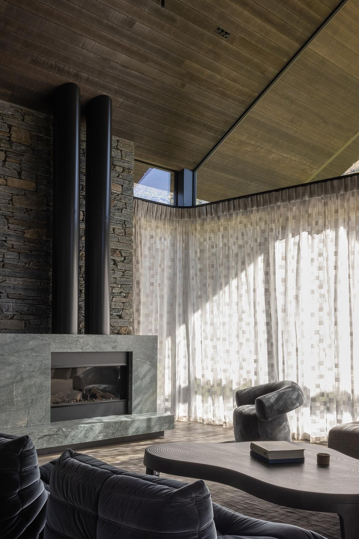 Soaring gable ceilings and expansive glazing flood the living room with natural light. Every angle has been considered to maximize the connection between indoors and the alpine panorama. The openness feels balanced by the grounding effect of the stone walls, giving the room both drama and warmth.