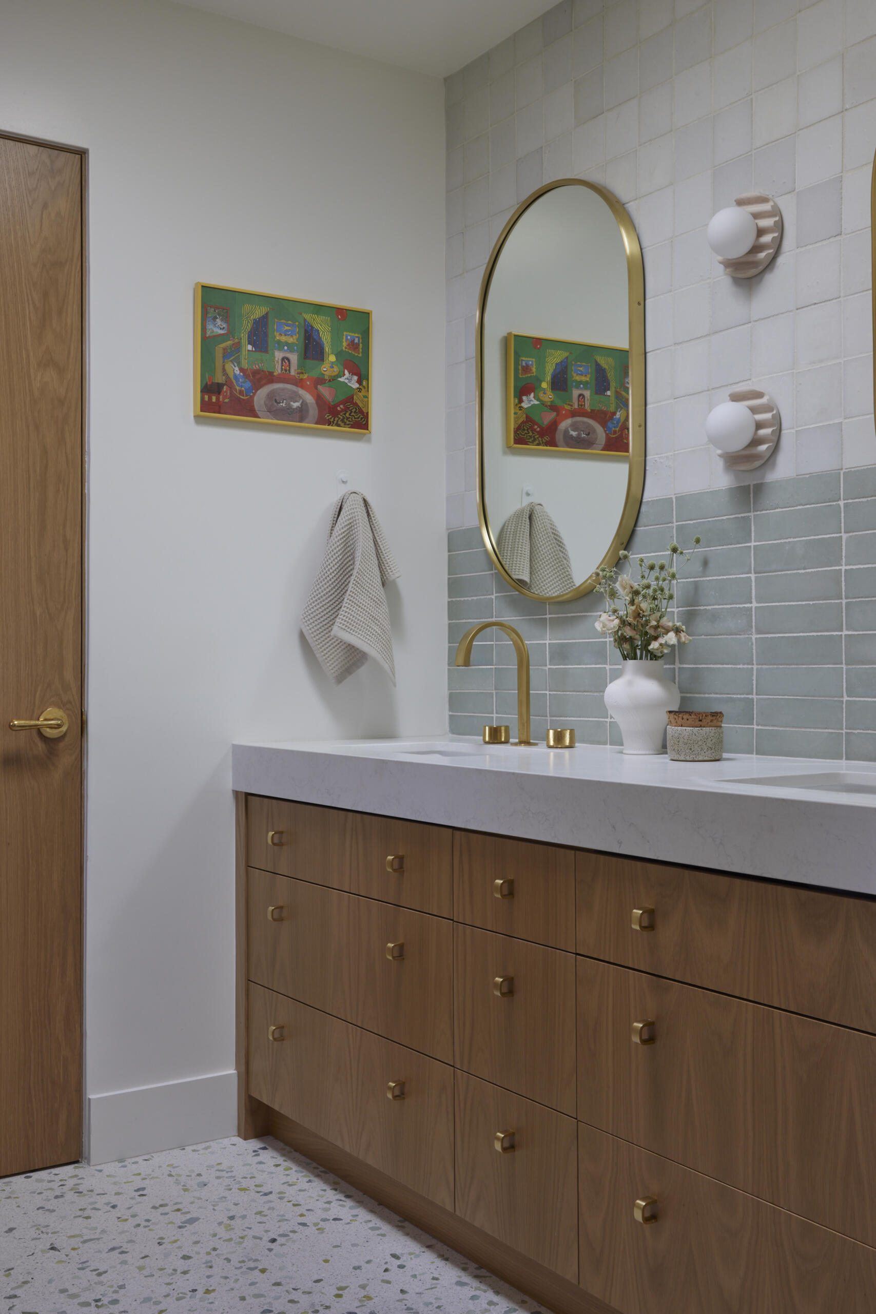 The kids bathroom strikes a balance between playful and practical. Terrazzo flooring provides a durable, easy-to-clean foundation, while soft green wall tiles add a calming touch. A wide vanity with wood drawers and brass hardware offers plenty of storage, designed to grow with the family. Rounded details, like the oval mirror, soften the clean lines, making the space inviting without losing its modern edge.
