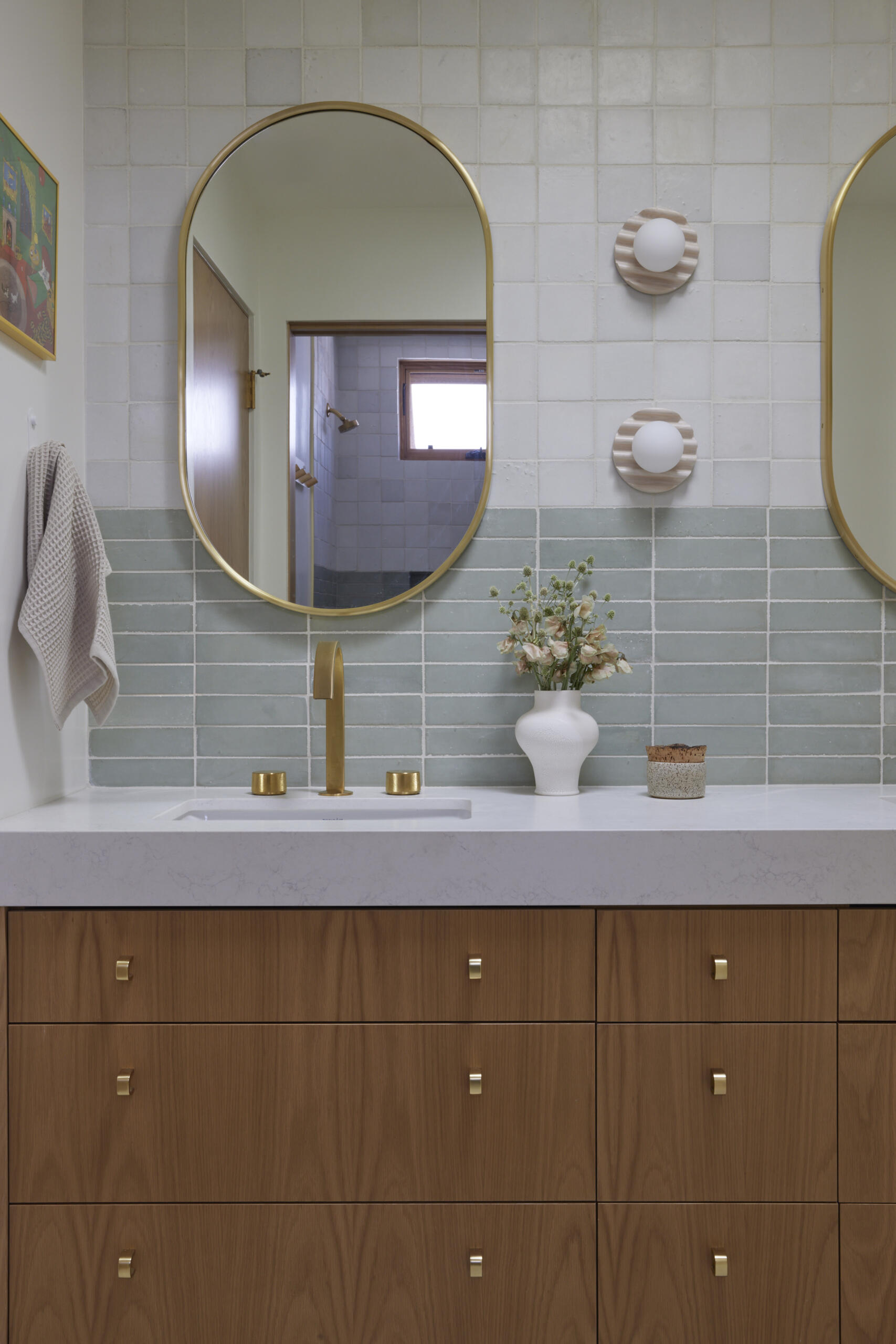 The kids bathroom strikes a balance between playful and practical. Terrazzo flooring provides a durable, easy-to-clean foundation, while soft green wall tiles add a calming touch. A wide vanity with wood drawers and brass hardware offers plenty of storage, designed to grow with the family. Rounded details, like the oval mirror, soften the clean lines, making the space inviting without losing its modern edge.