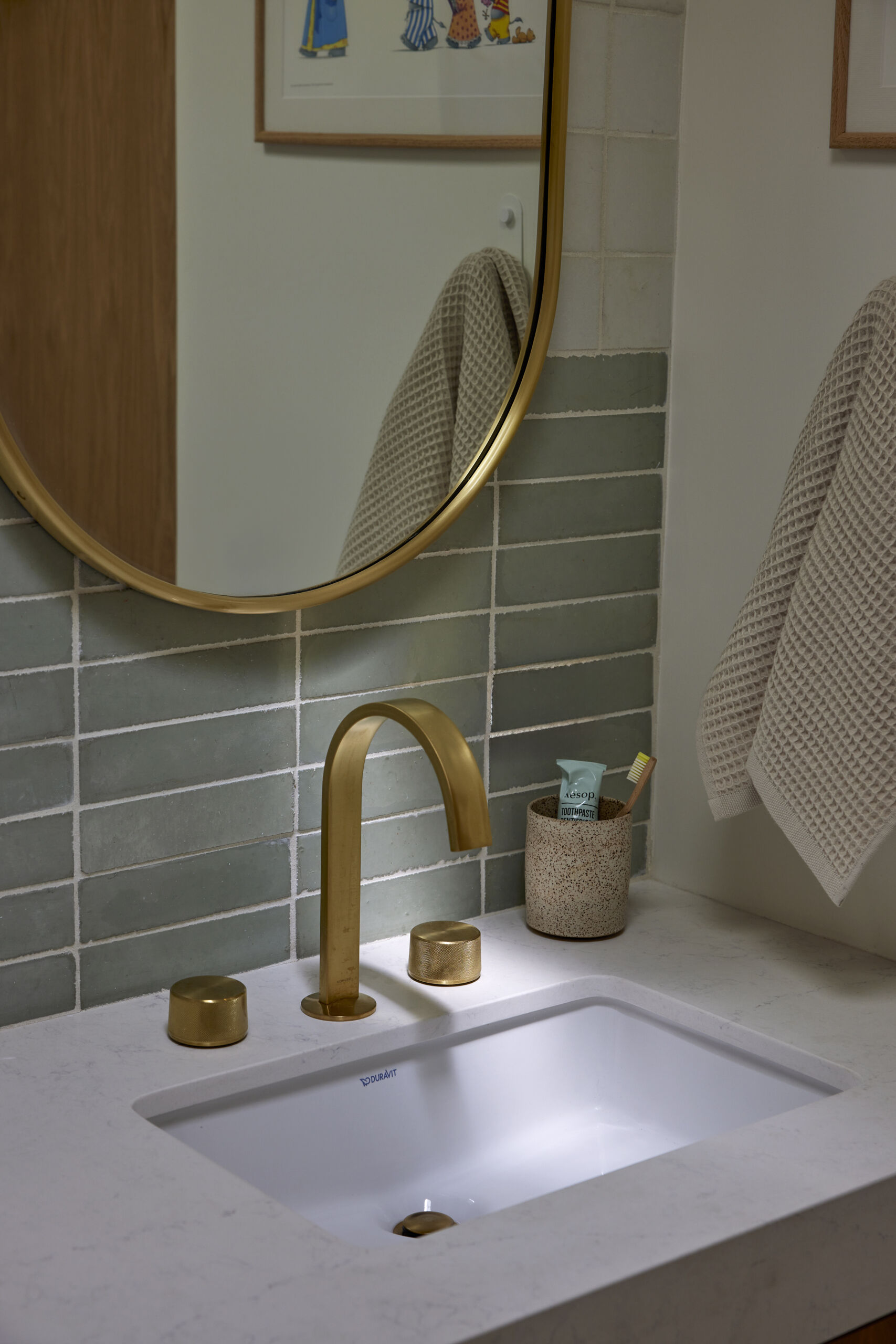 The kids bathroom strikes a balance between playful and practical. Terrazzo flooring provides a durable, easy-to-clean foundation, while soft green wall tiles add a calming touch. A wide vanity with wood drawers and brass hardware offers plenty of storage, designed to grow with the family. Rounded details, like the oval mirror, soften the clean lines, making the space inviting without losing its modern edge.