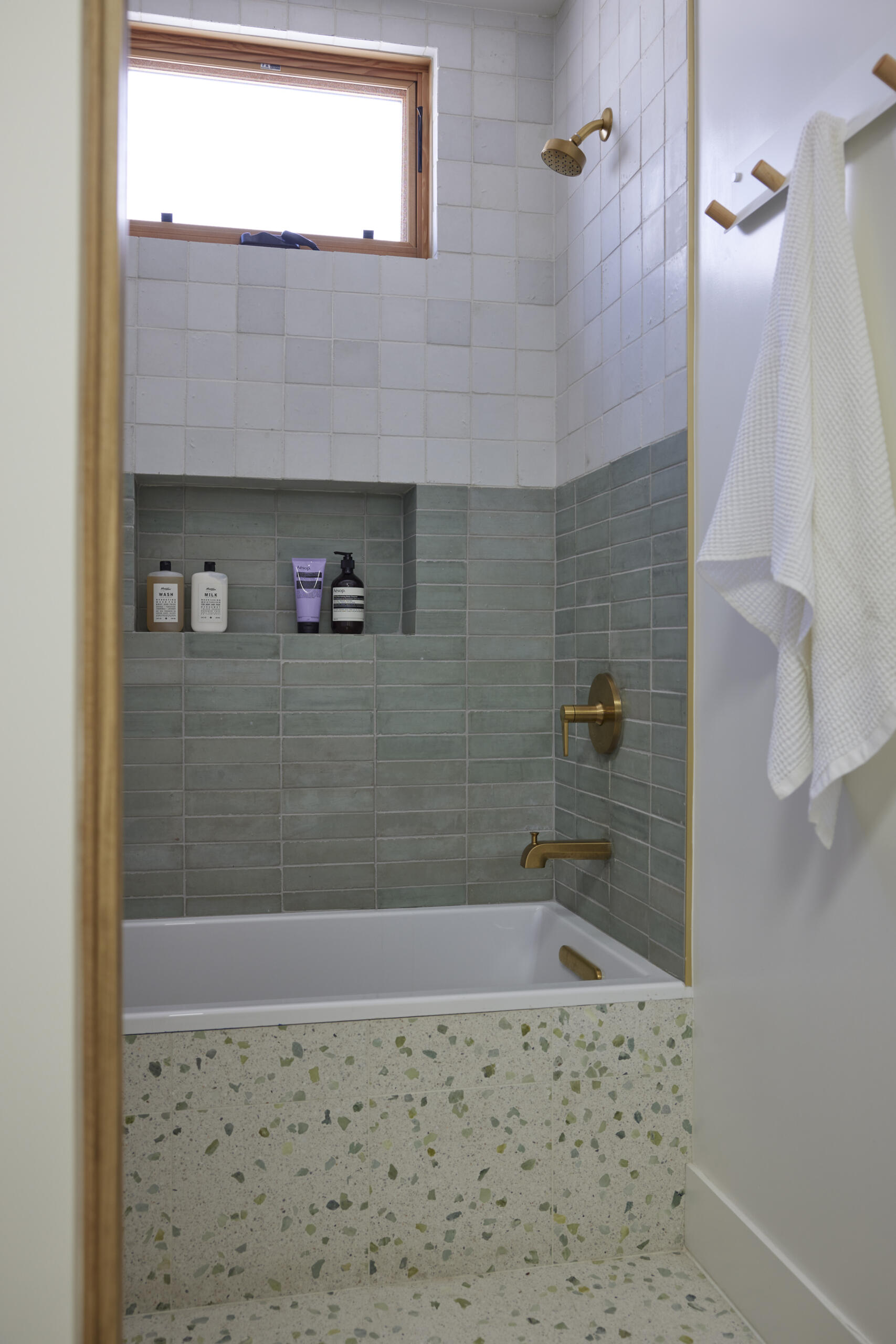 The kids bathroom strikes a balance between playful and practical. Terrazzo flooring provides a durable, easy-to-clean foundation, while soft green wall tiles add a calming touch. A wide vanity with wood drawers and brass hardware offers plenty of storage, designed to grow with the family. Rounded details, like the oval mirror, soften the clean lines, making the space inviting without losing its modern edge.