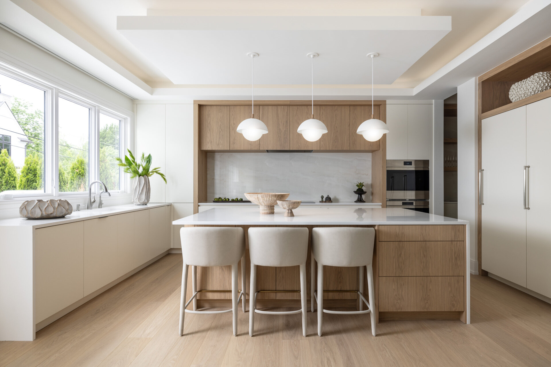 In the kitchen, wood and white do the talking. Three sculptural globe pendants hover above a generous island, while quiet cabinetry keeps the visual noise to a minimum. The palette is restrained, the textures do the lifting, and the result is exactly what busy mornings need. Practical yet polished. Appliances tuck away. Traffic flows. The whole space feels calm and collected.
