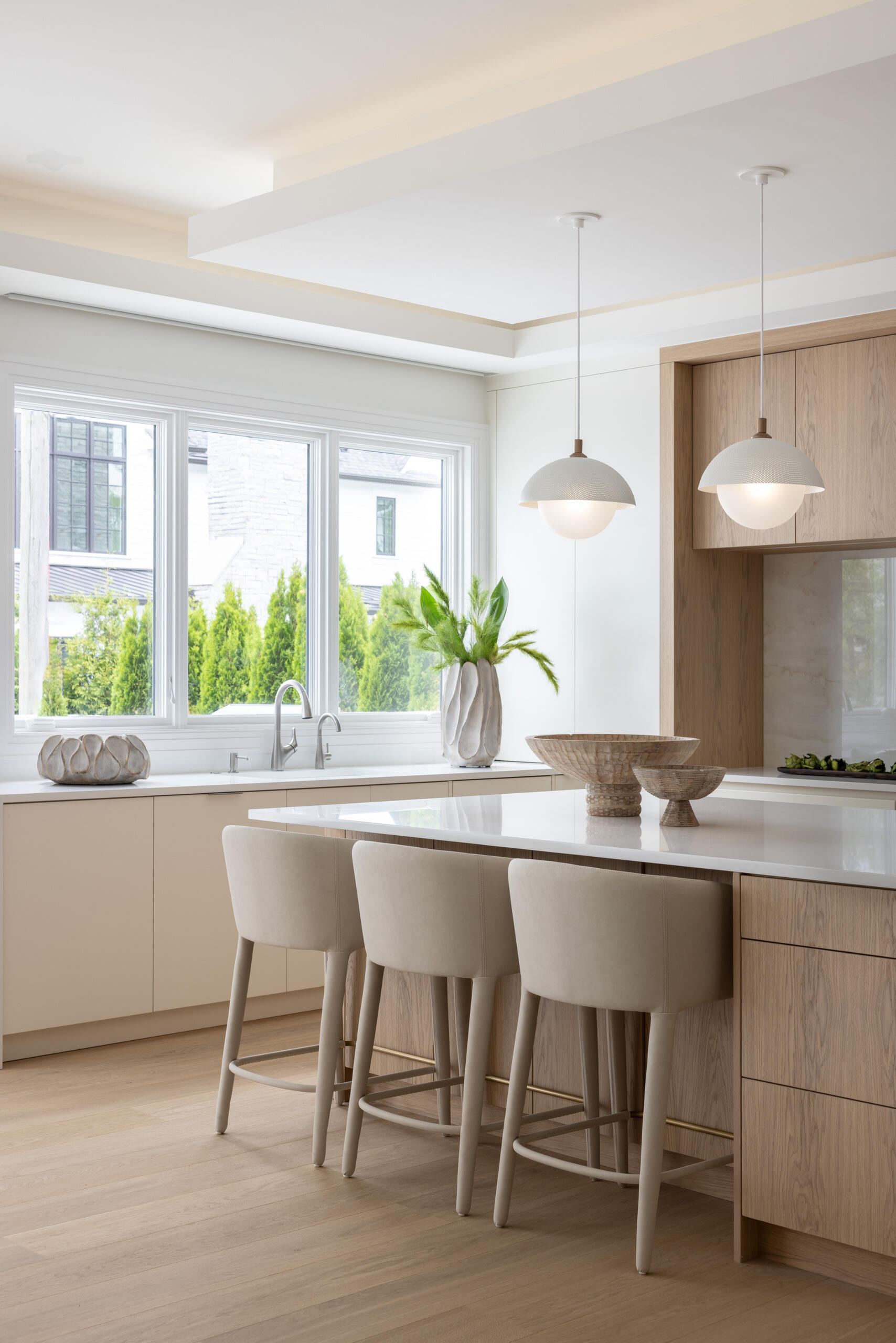In the kitchen, wood and white do the talking. Three sculptural globe pendants hover above a generous island, while quiet cabinetry keeps the visual noise to a minimum. The palette is restrained, the textures do the lifting, and the result is exactly what busy mornings need. Practical yet polished. Appliances tuck away. Traffic flows. The whole space feels calm and collected.