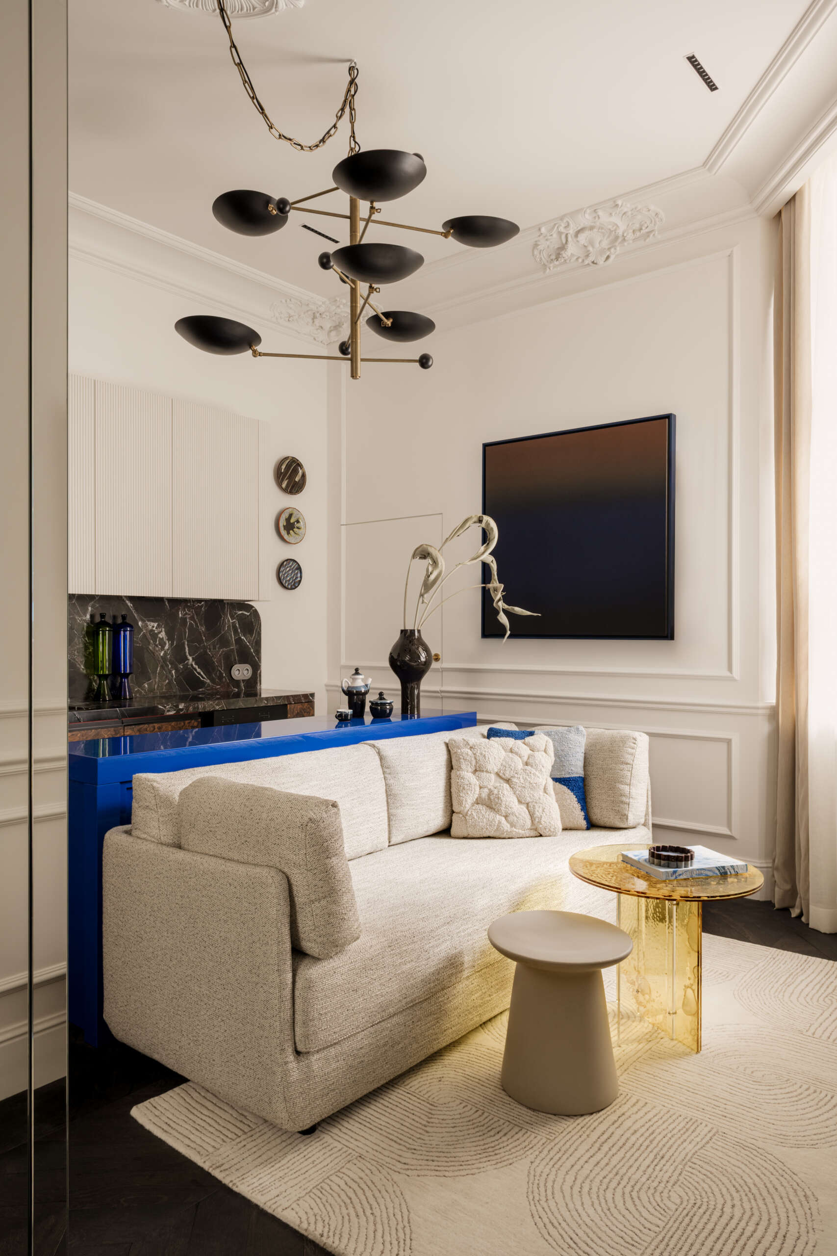 The living area behaves like a miniature salon, with layered art, a vintage brass chandelier, and a bold yellow plexiglass table. A deep sofa and curated Polish design pieces turn the compact footprint into a scene of effortless charm.