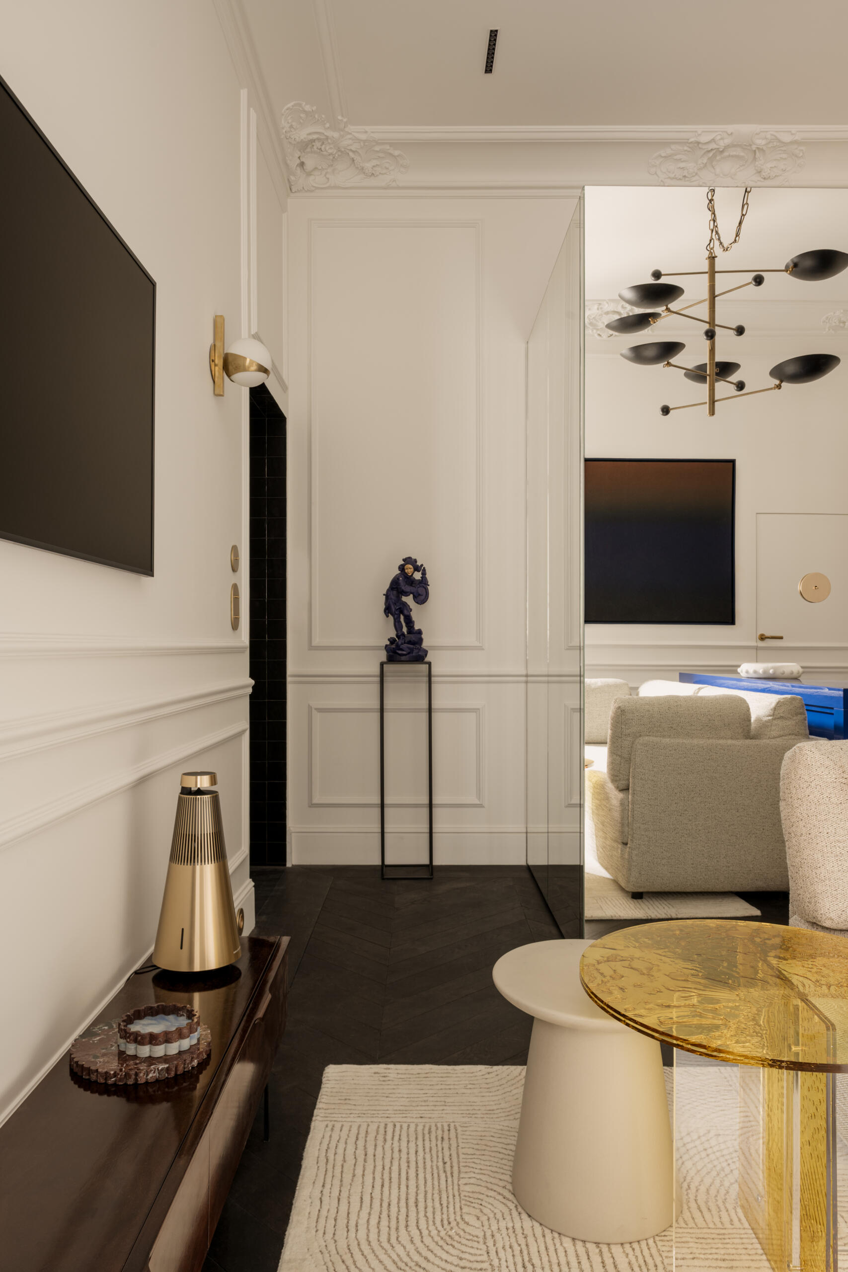 The living area behaves like a miniature salon, with layered art, a vintage brass chandelier, and a bold yellow plexiglass table. A deep sofa and curated Polish design pieces turn the compact footprint into a scene of effortless charm.