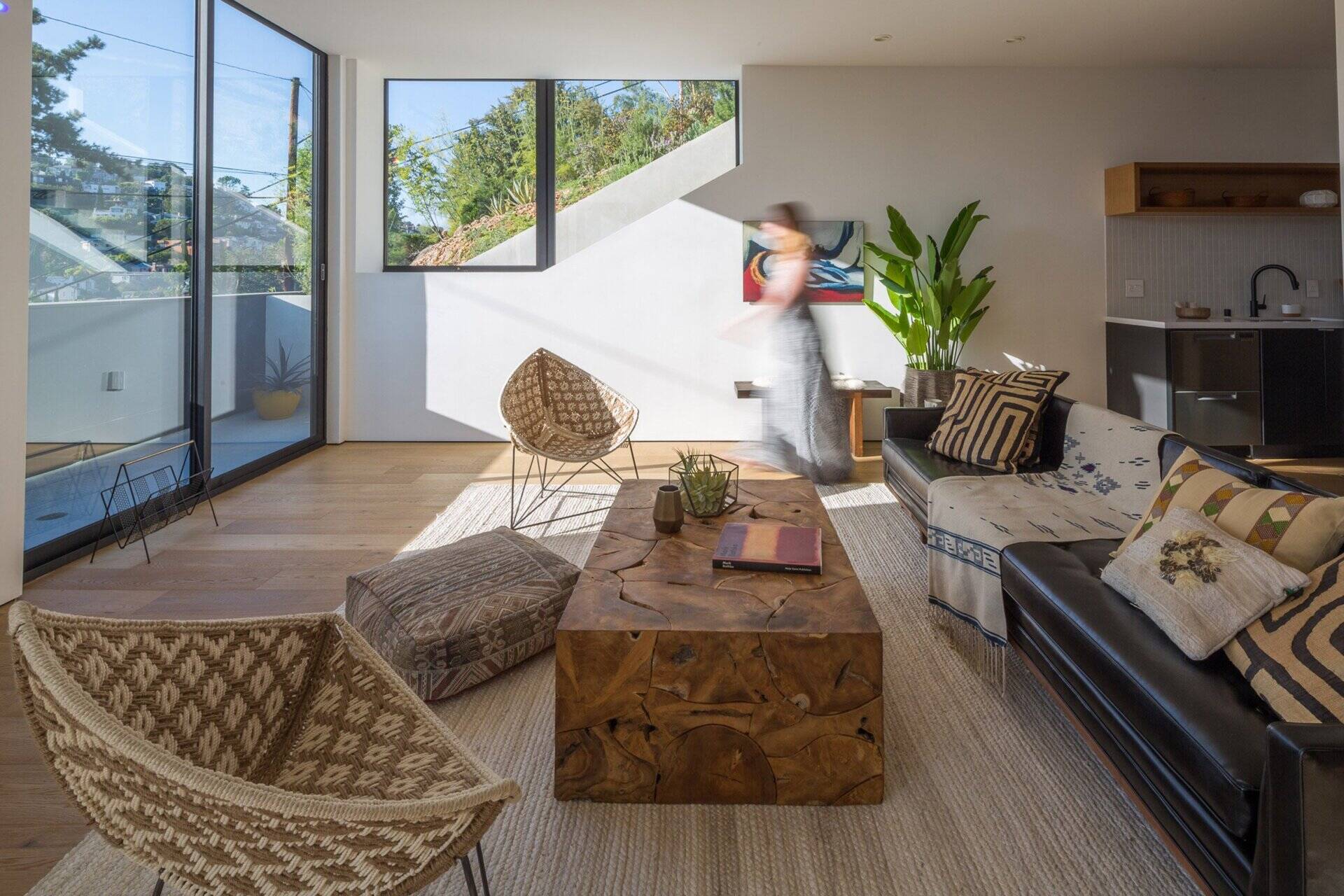 Inside, the open-plan living room, dining area, and kitchen unfold across the main floor. Large windows and sliding glass doors dissolve the line between inside and out, opening to a small deck with seating that extends the living area into the hillside.