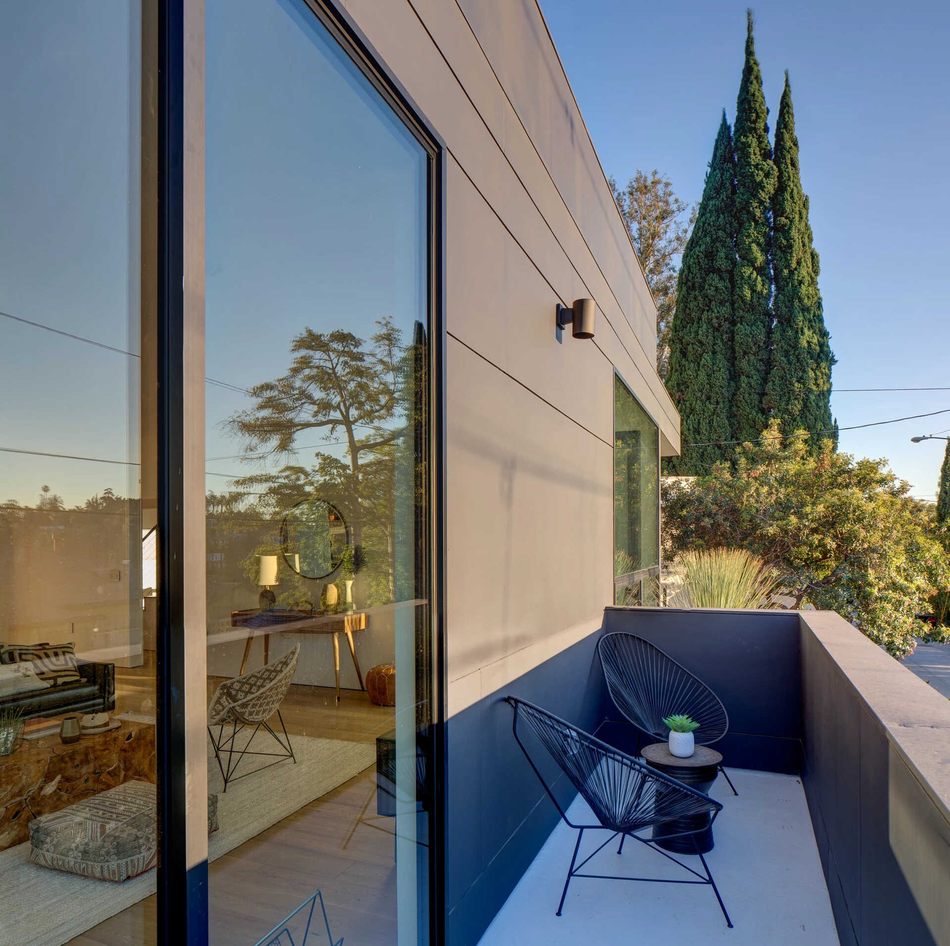 Inside, the open-plan living room, dining area, and kitchen unfold across the main floor. Large windows and sliding glass doors dissolve the line between inside and out, opening to a small deck with seating that extends the living area into the hillside.