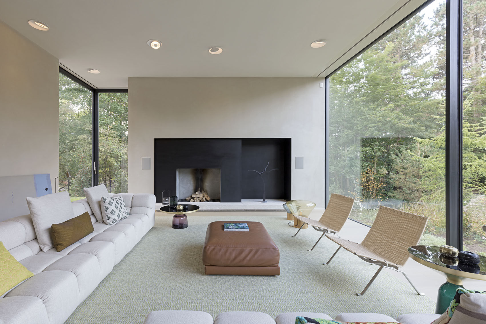 In the living room, a dark fireplace anchors the far wall, its tone a subtle echo of the home’s black facade. Vast windows frame the surrounding landscape, while floor-to-ceiling glass along the kitchen and living areas allows daylight to flood the space.