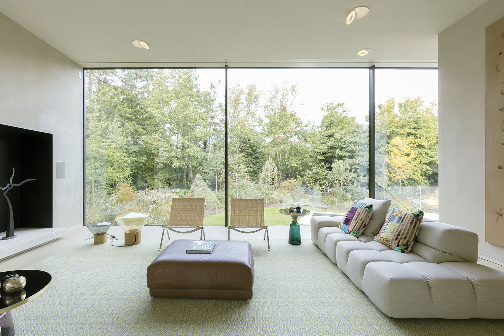 In the living room, a dark fireplace anchors the far wall, its tone a subtle echo of the home’s black facade. Vast windows frame the surrounding landscape, while floor-to-ceiling glass along the kitchen and living areas allows daylight to flood the space.