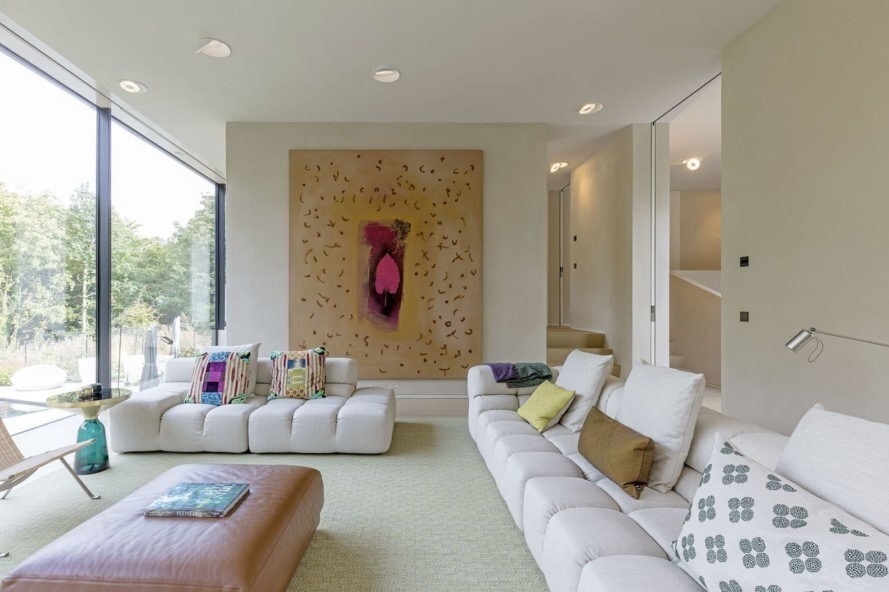 In the living room, a dark fireplace anchors the far wall, its tone a subtle echo of the home’s black facade. Vast windows frame the surrounding landscape, while floor-to-ceiling glass along the kitchen and living areas allows daylight to flood the space.