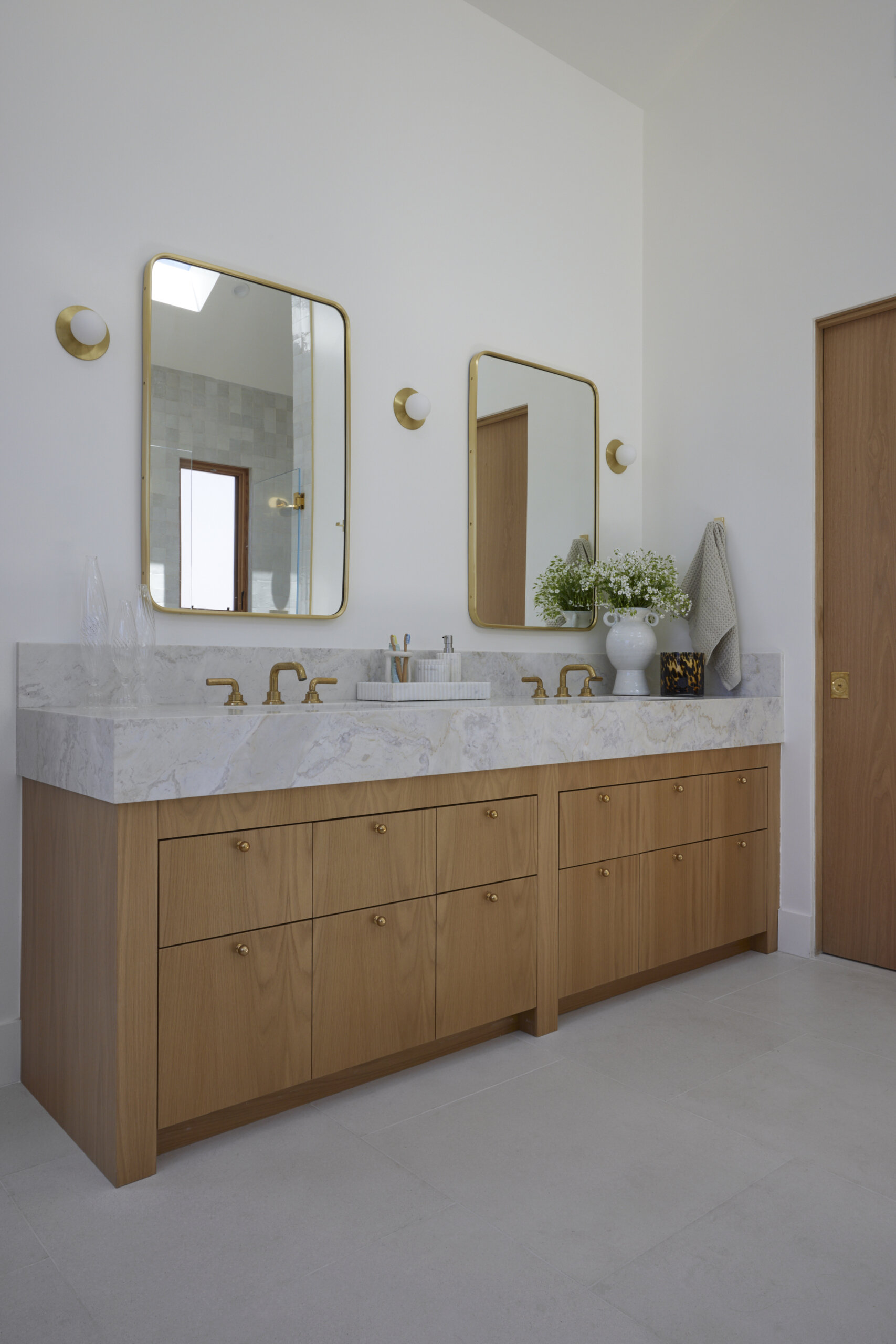 The primary bath feels fresh, refined, and serene, where pale wood cabinetry adds warmth beneath a crisp marble countertop. Soft natural light filters through frosted windows, illuminating the veined marble framing a deep soaking tub and the walk-in shower lined with pale square tiles, each catching the light differently. Brass faucets, hardware, slim-framed mirrors, and wall sconces introduce a soft golden glow against the clean white walls, keeping the space open and airy.
