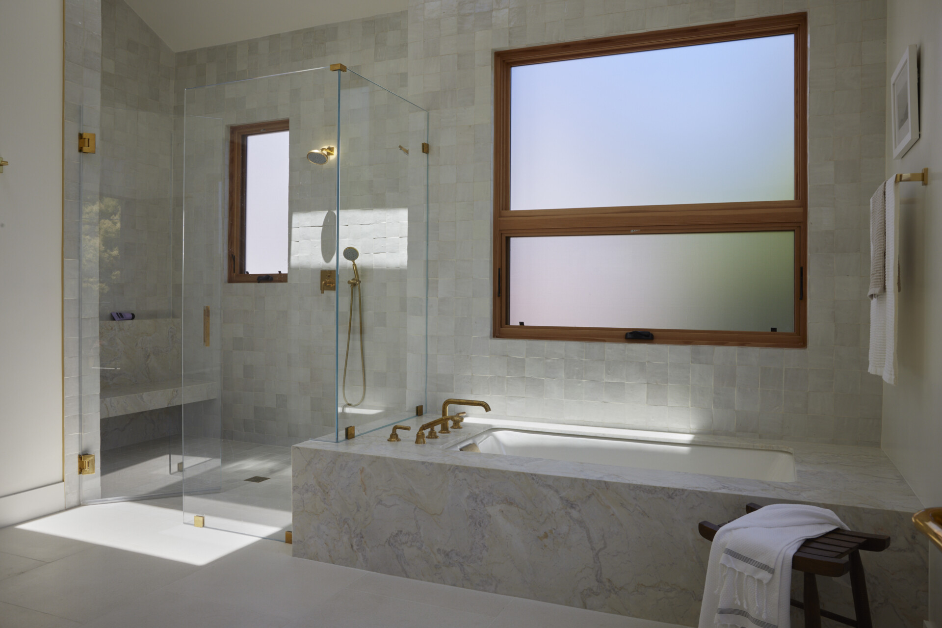 The primary bath feels fresh, refined, and serene, where pale wood cabinetry adds warmth beneath a crisp marble countertop. Soft natural light filters through frosted windows, illuminating the veined marble framing a deep soaking tub and the walk-in shower lined with pale square tiles, each catching the light differently. Brass faucets, hardware, slim-framed mirrors, and wall sconces introduce a soft golden glow against the clean white walls, keeping the space open and airy.