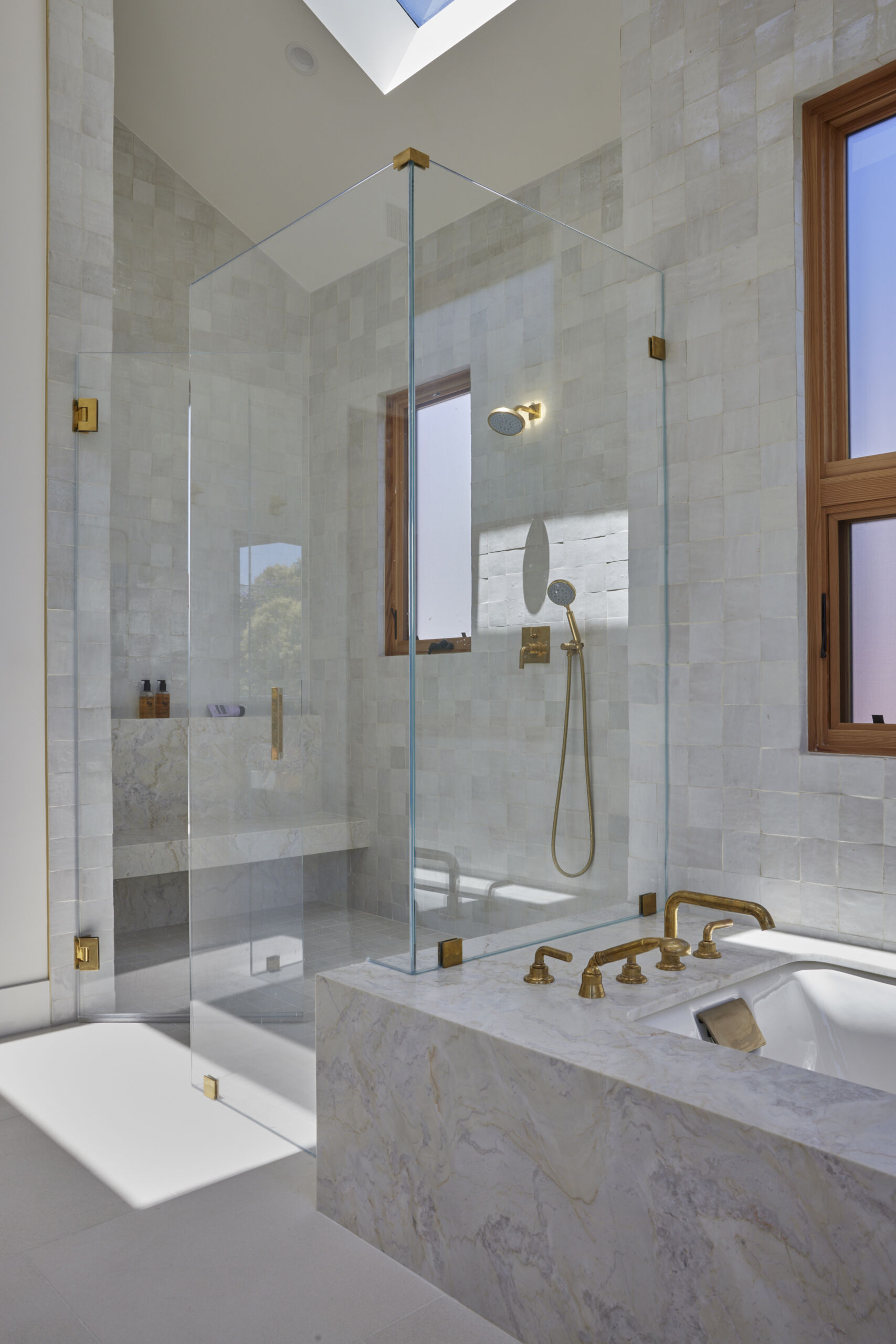 The primary bath feels fresh, refined, and serene, where pale wood cabinetry adds warmth beneath a crisp marble countertop. Soft natural light filters through frosted windows, illuminating the veined marble framing a deep soaking tub and the walk-in shower lined with pale square tiles, each catching the light differently. Brass faucets, hardware, slim-framed mirrors, and wall sconces introduce a soft golden glow against the clean white walls, keeping the space open and airy.