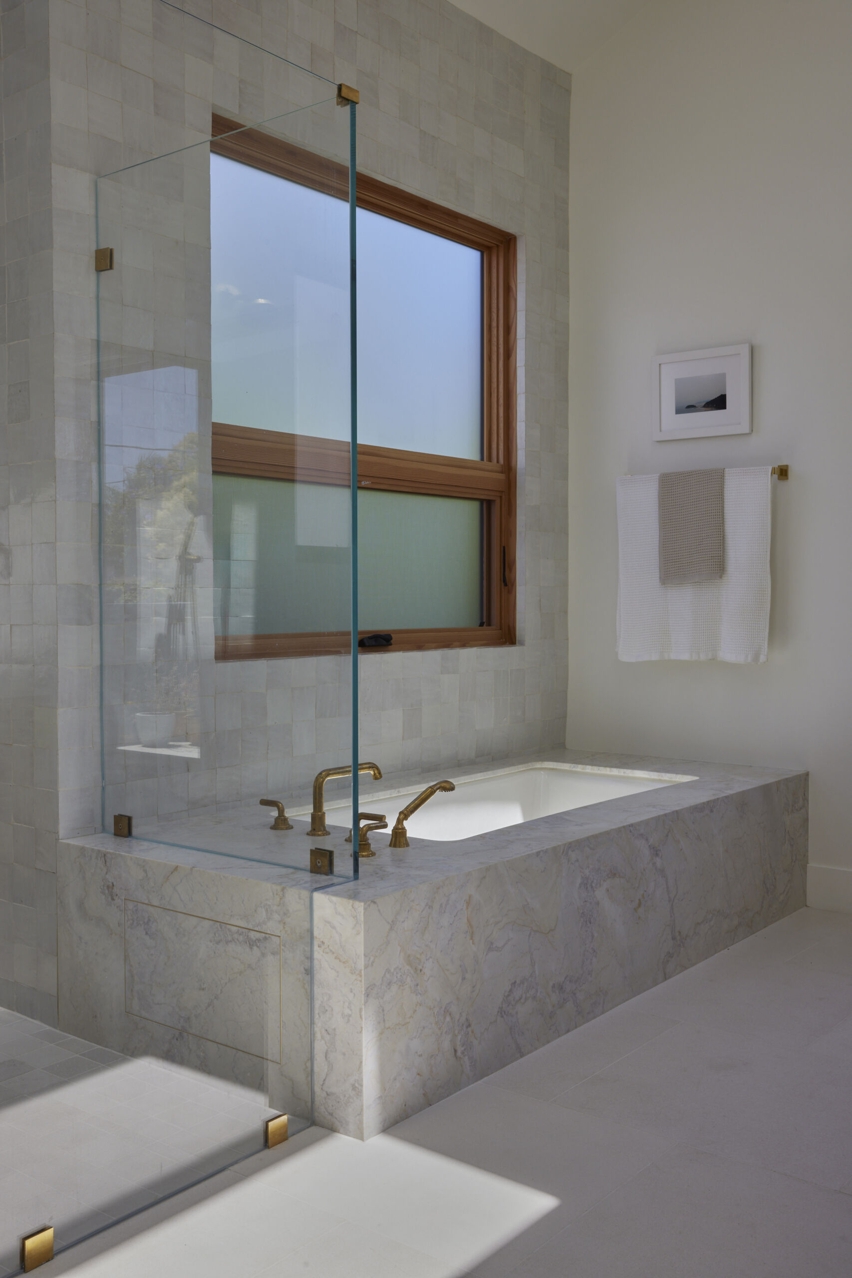 The primary bath feels fresh, refined, and serene, where pale wood cabinetry adds warmth beneath a crisp marble countertop. Soft natural light filters through frosted windows, illuminating the veined marble framing a deep soaking tub and the walk-in shower lined with pale square tiles, each catching the light differently. Brass faucets, hardware, slim-framed mirrors, and wall sconces introduce a soft golden glow against the clean white walls, keeping the space open and airy.