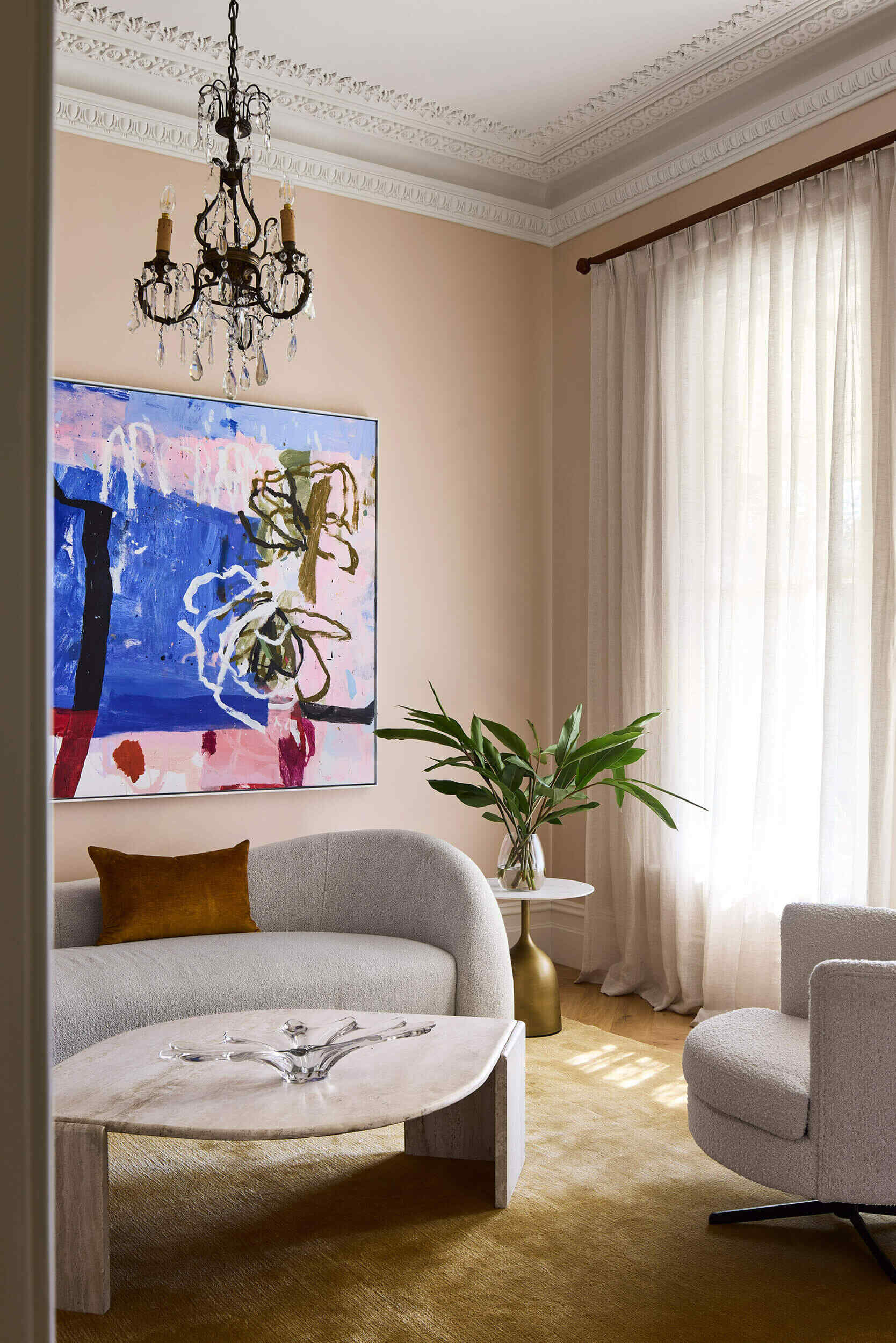 At the front of this home, the sitting room captures the spirit of the original Victorian architecture while embracing a calm, contemporary sensibility. Ornate ceiling cornices and a crystal chandelier speak to the home’s history, yet the palette and furnishings introduce softness and modern restraint.