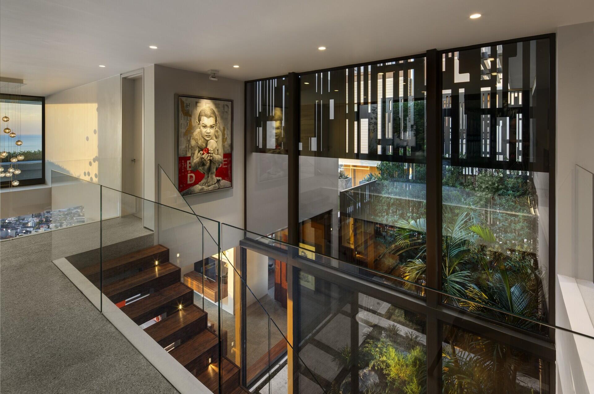 Wood and black metal stairs, paired with invisible glass railings, lead to the upper floor, where a landing overlooks both the living room and courtyard.