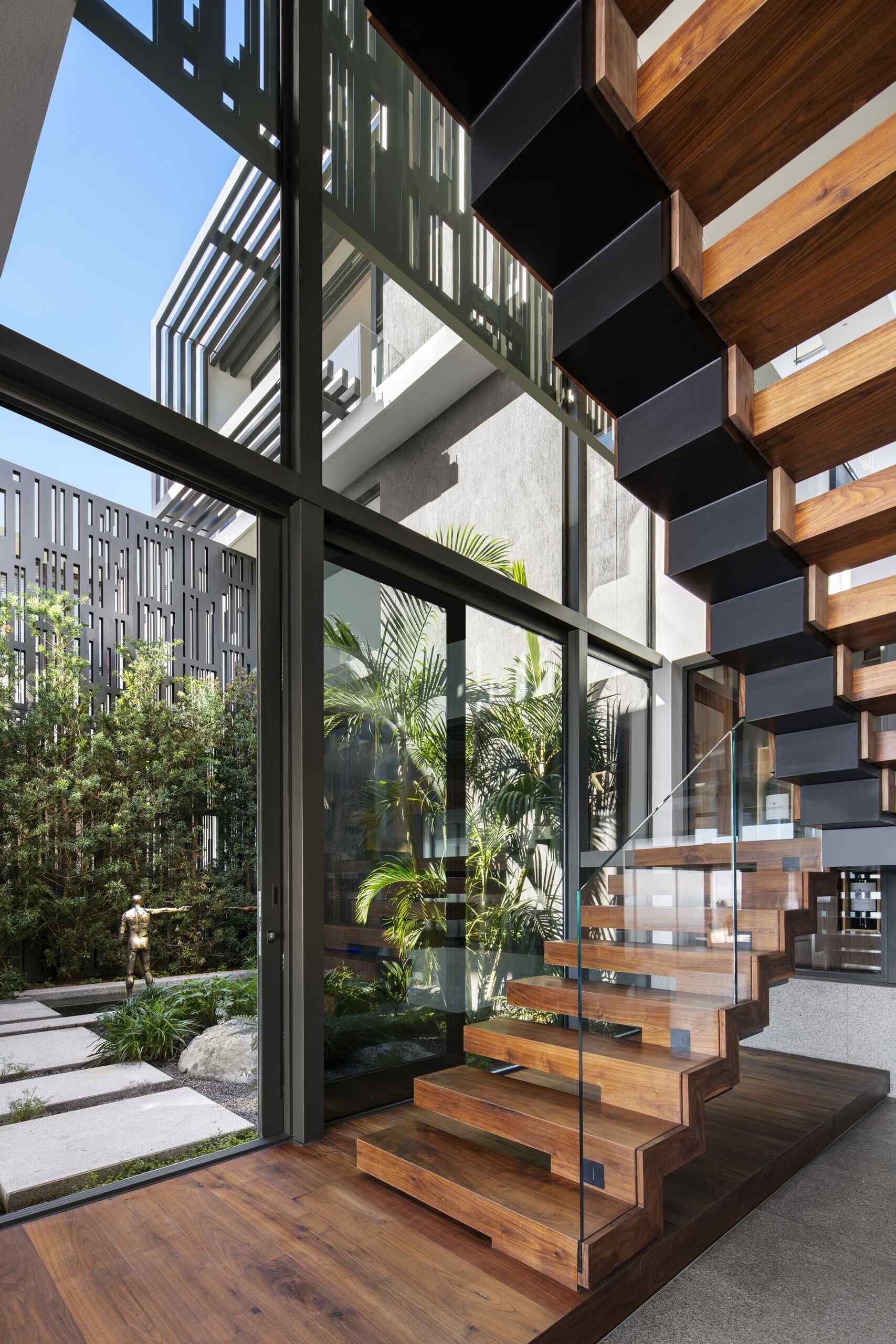 Wood and black metal stairs, paired with invisible glass railings, lead to the upper floor, where a landing overlooks both the living room and courtyard.