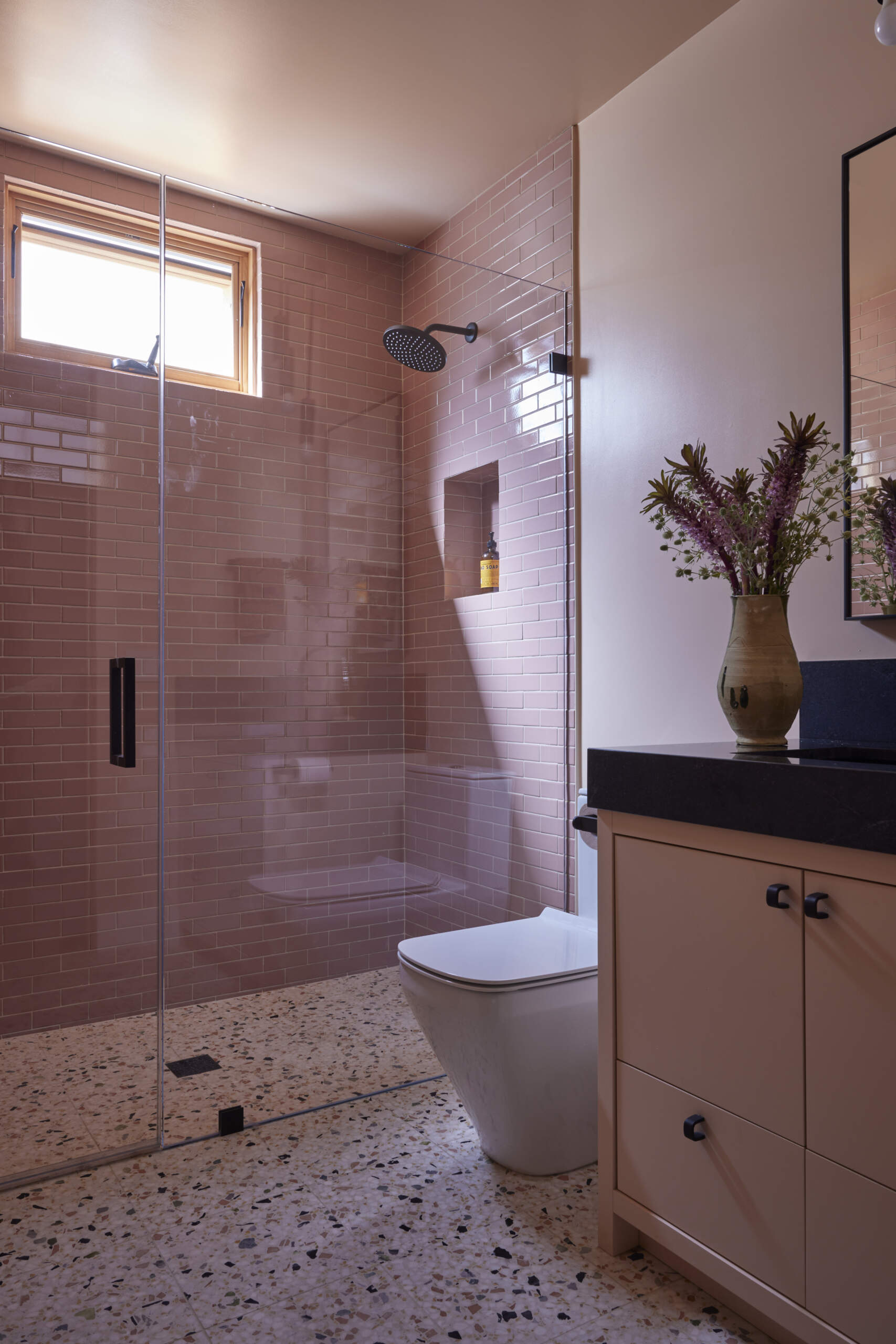 The guest bath carries thoughtful details and a soft pop of color with pink tiles and walls, while terrazzo flooring has a curbless transition and the shower niches combine function with precision.