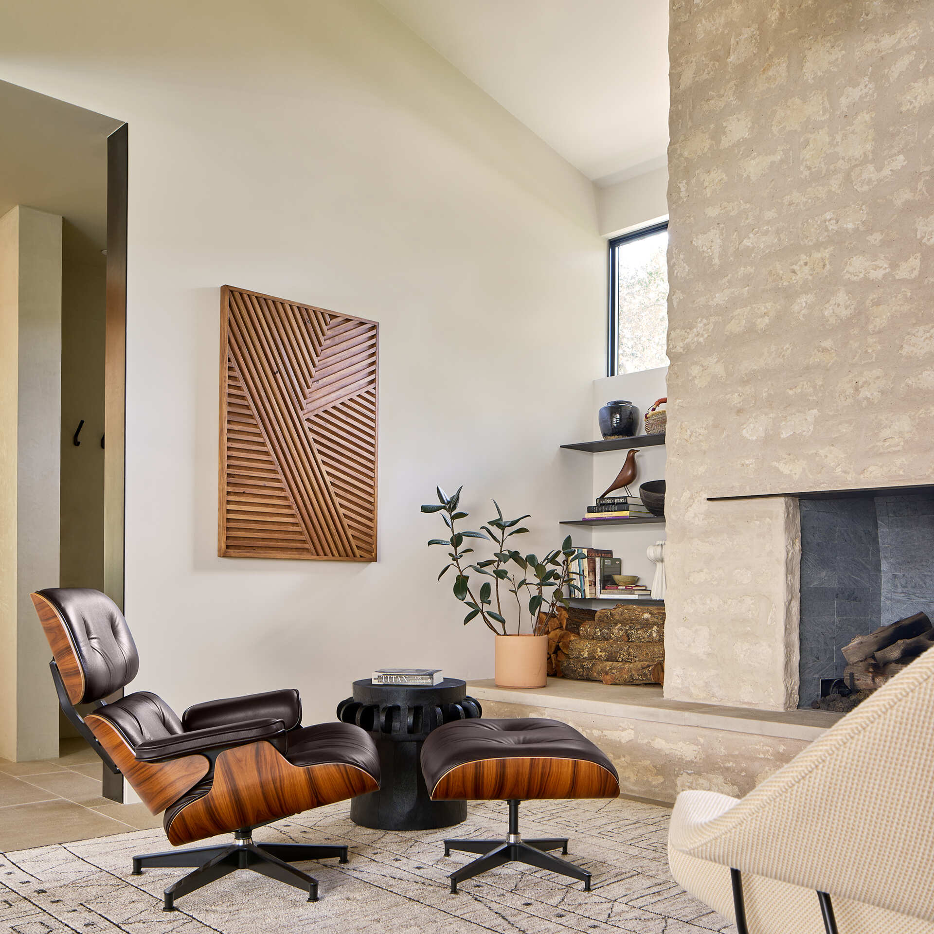Anchored by a textured stone fireplace, this smaller sitting room blends natural materials with mid-century modern design. An iconic Eames lounge chair and ottoman in warm wood and leather invite relaxation, paired with a sculptural black side table that adds contrast, while built-in shelving holds books, ceramics, and art objects, giving the room a curated, personal feel.
