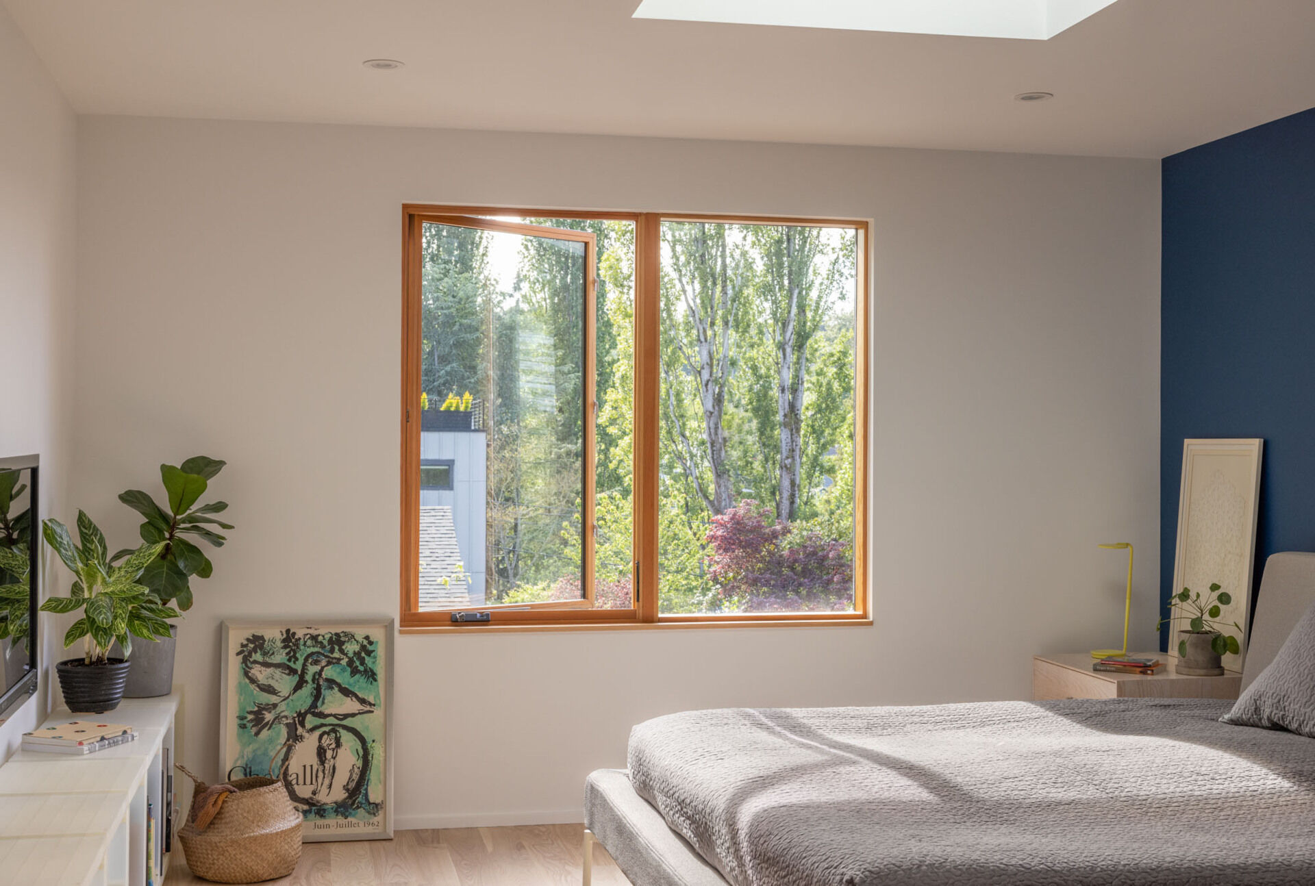 The primary suite was reshaped for privacy and oriented toward views of mature poplars. New ribbon windows frame the foliage, creating a serene backdrop, while a blue accent wall adds color to the room. 