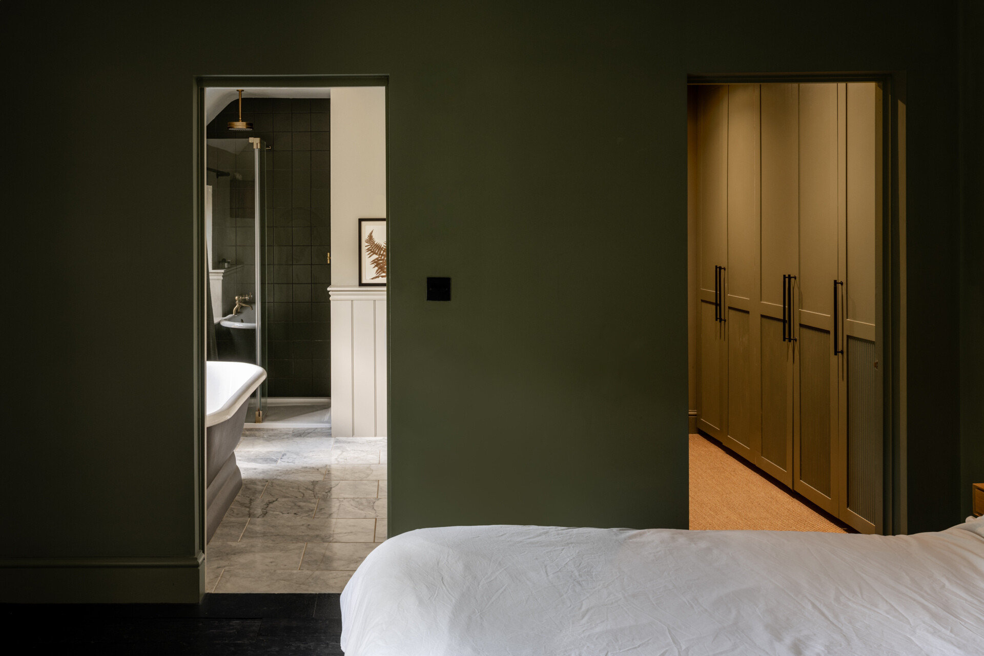 A calm bedroom painted in rich olive tones opens to a marble floored bathroom with a classic tub and dark tiled shower, while a warm walk-in wardrobe glows softly to the side. The layout feels intentional and serene, with each space connected by subtle light and thoughtful detailing.